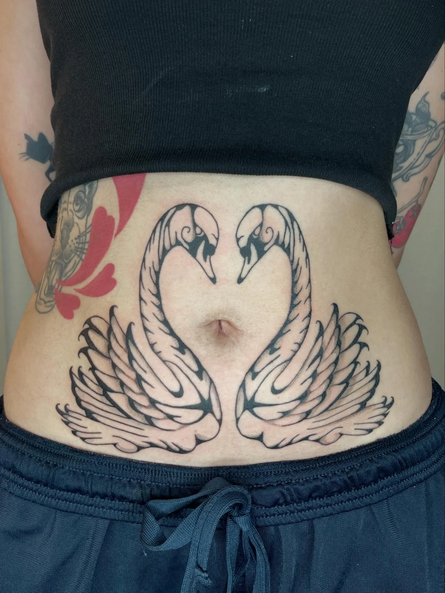 Tattoo of a symmetrical swan design on a person's lower abdomen, with tattoos on both arms visible in the background.