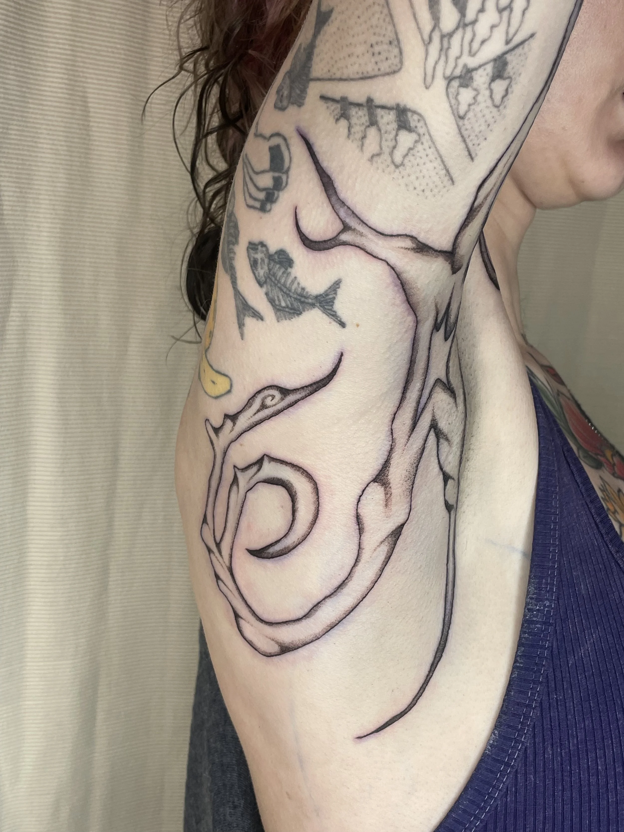 A person's upper arm with various black and grey tattoos including a fish, a whale, a star, and a boat, with a part of the face and shoulder visible.