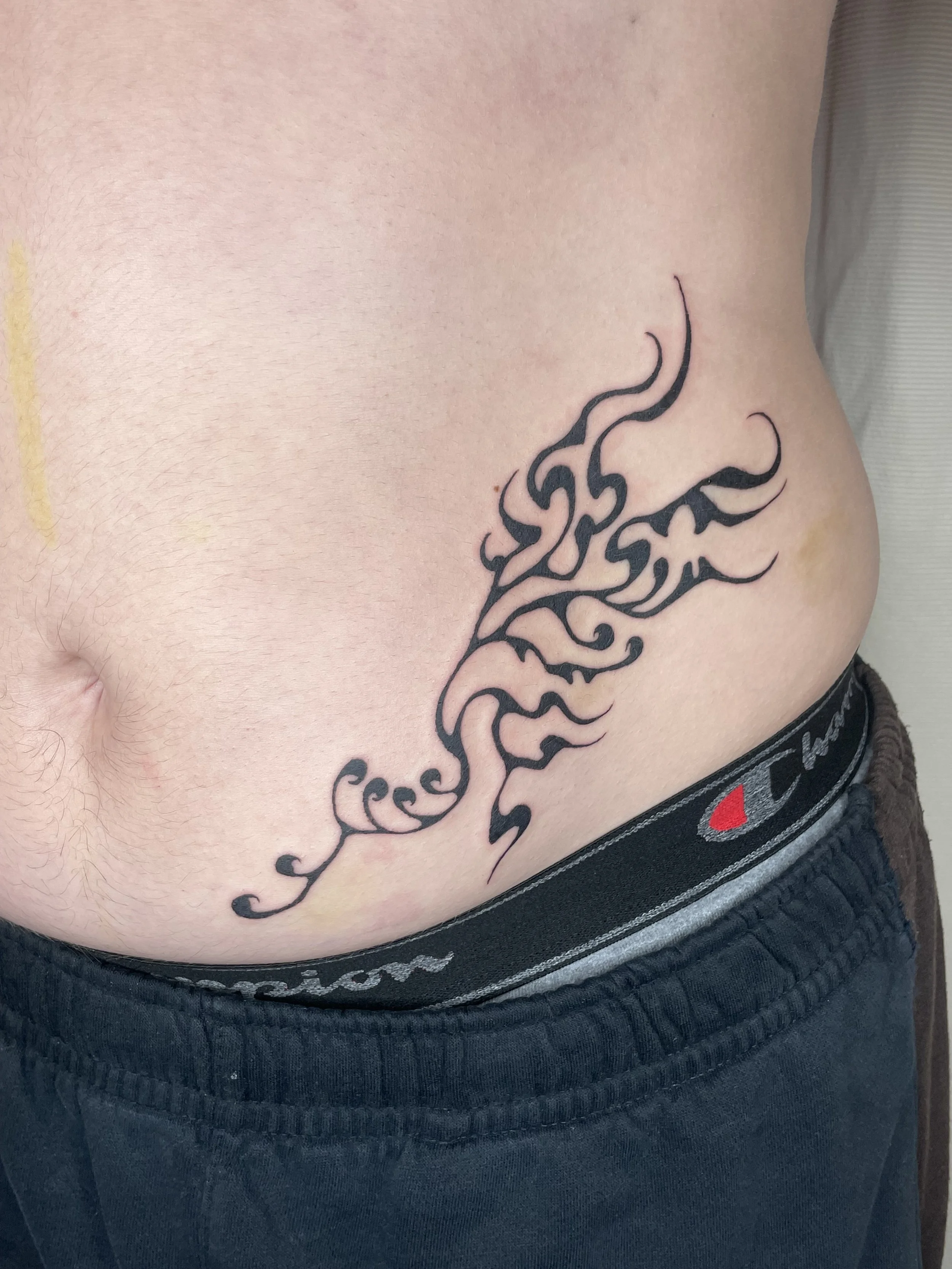 Close-up of a person's side with a black tribal-style tattoo on their rib cage, partially visible above the waistband of dark boxer briefs.