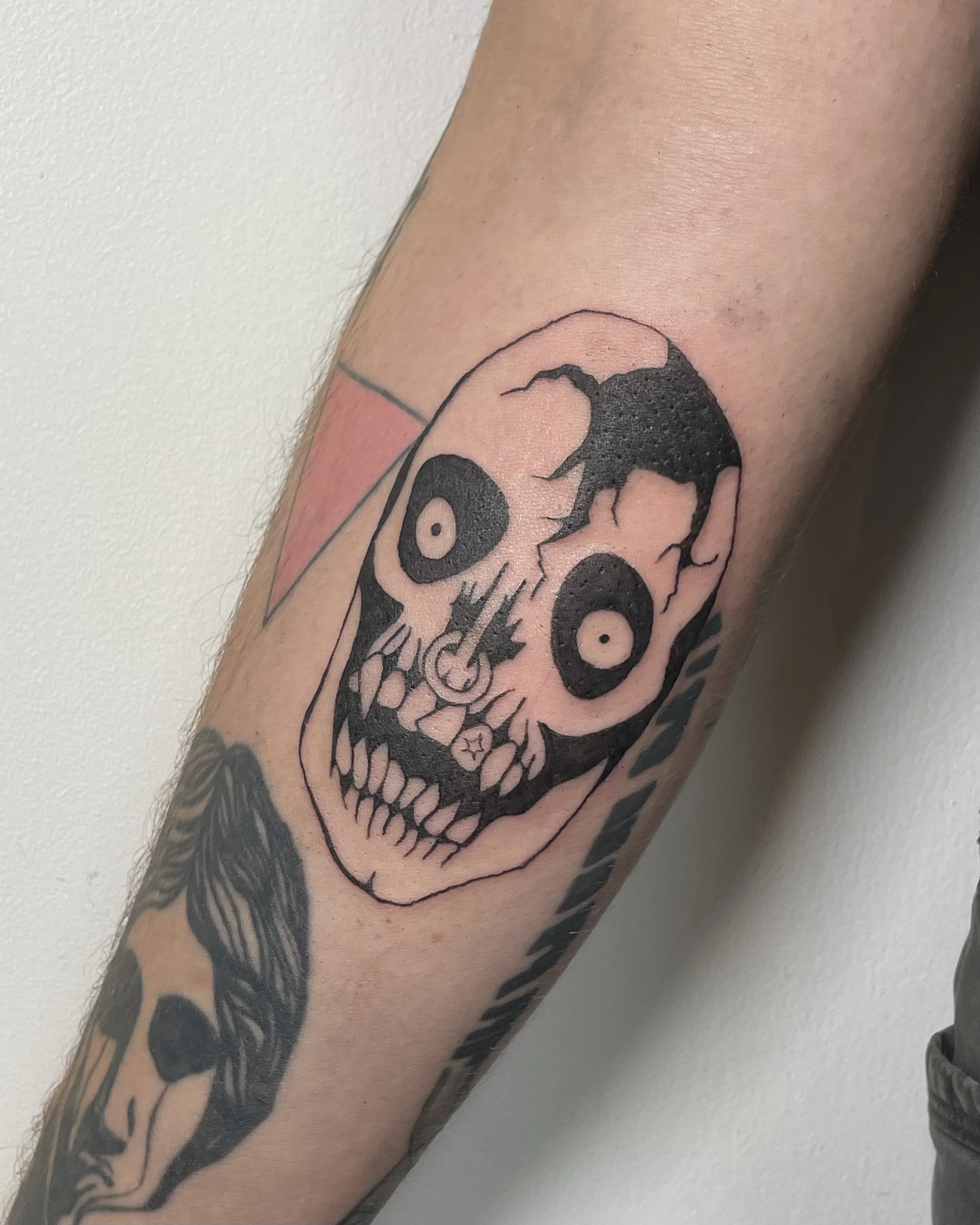 Tattoo of a stylized skull with large round eyes and cracked features on a person's arm.