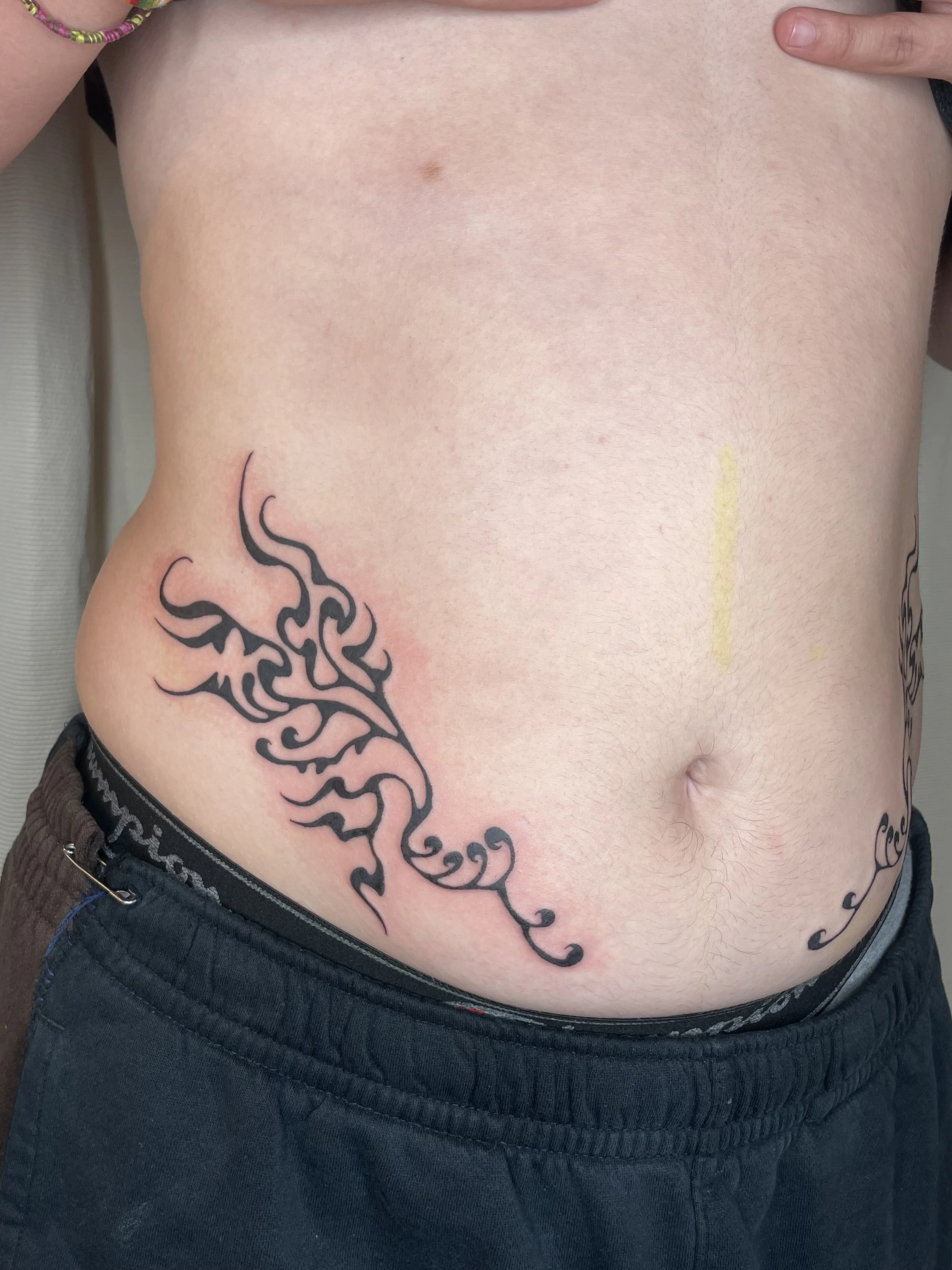 A person's torso revealing a tattoo of black tribal design on the left side of the ribcage near the waist. The person is wearing dark pants with a visible belt.