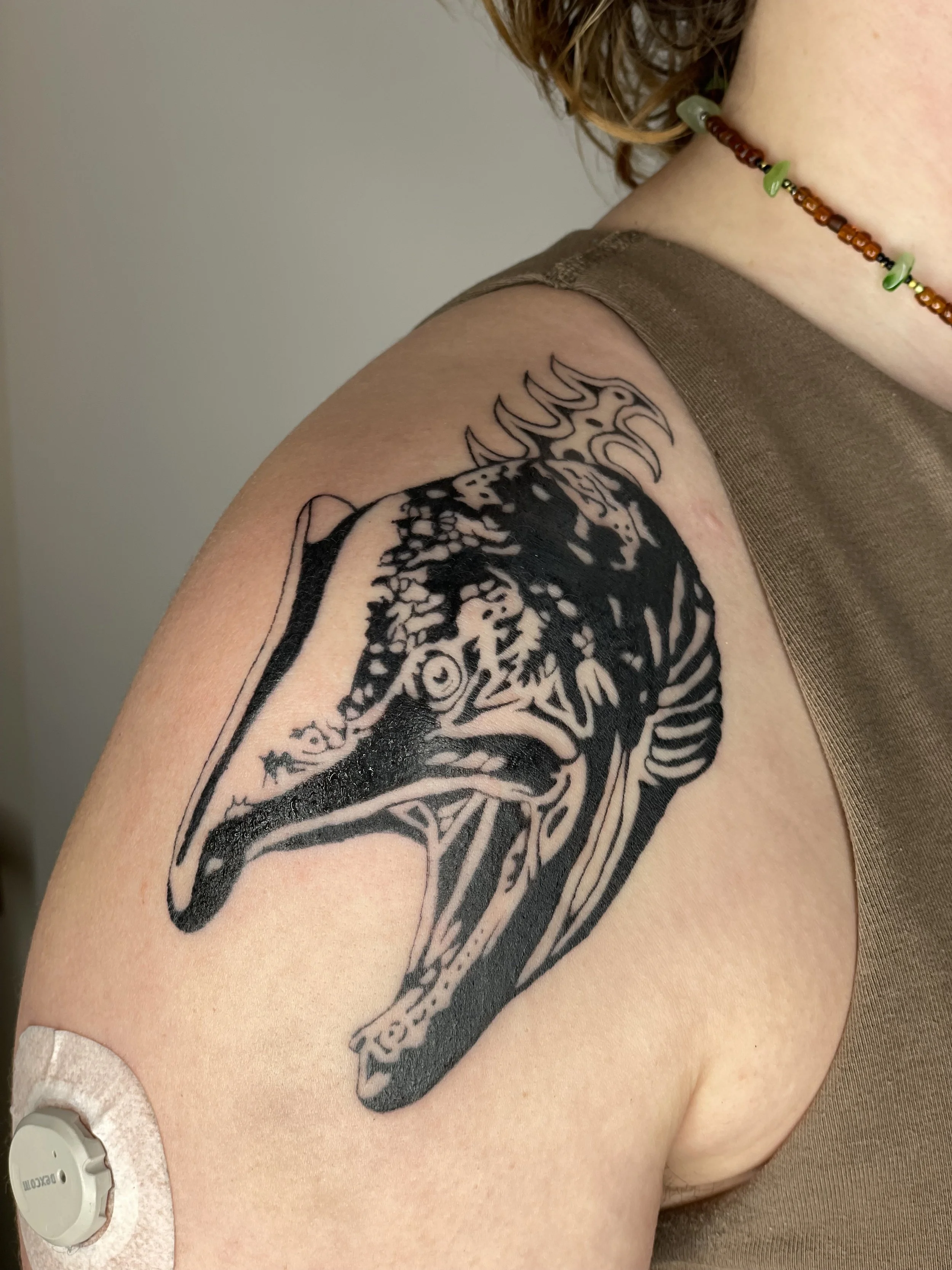 Close-up of a person's upper arm with a black and grey tattoo of a stylized salmon along the shoulder. The person is wearing a brown sleeveless top and a beaded necklace with green and brown beads.