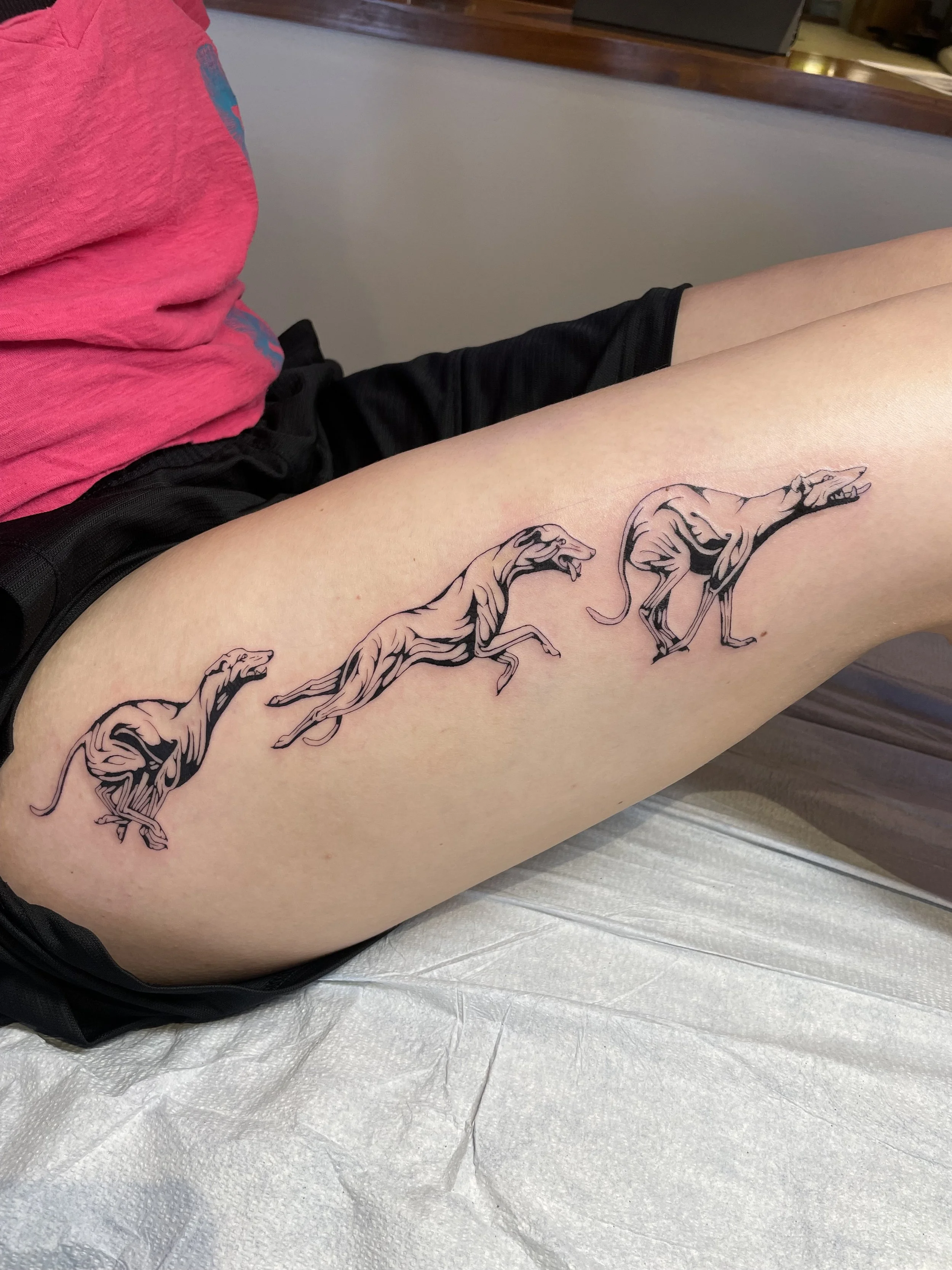 Tattoo of a pack greyhounds on the thigh of a person who is sitting down.