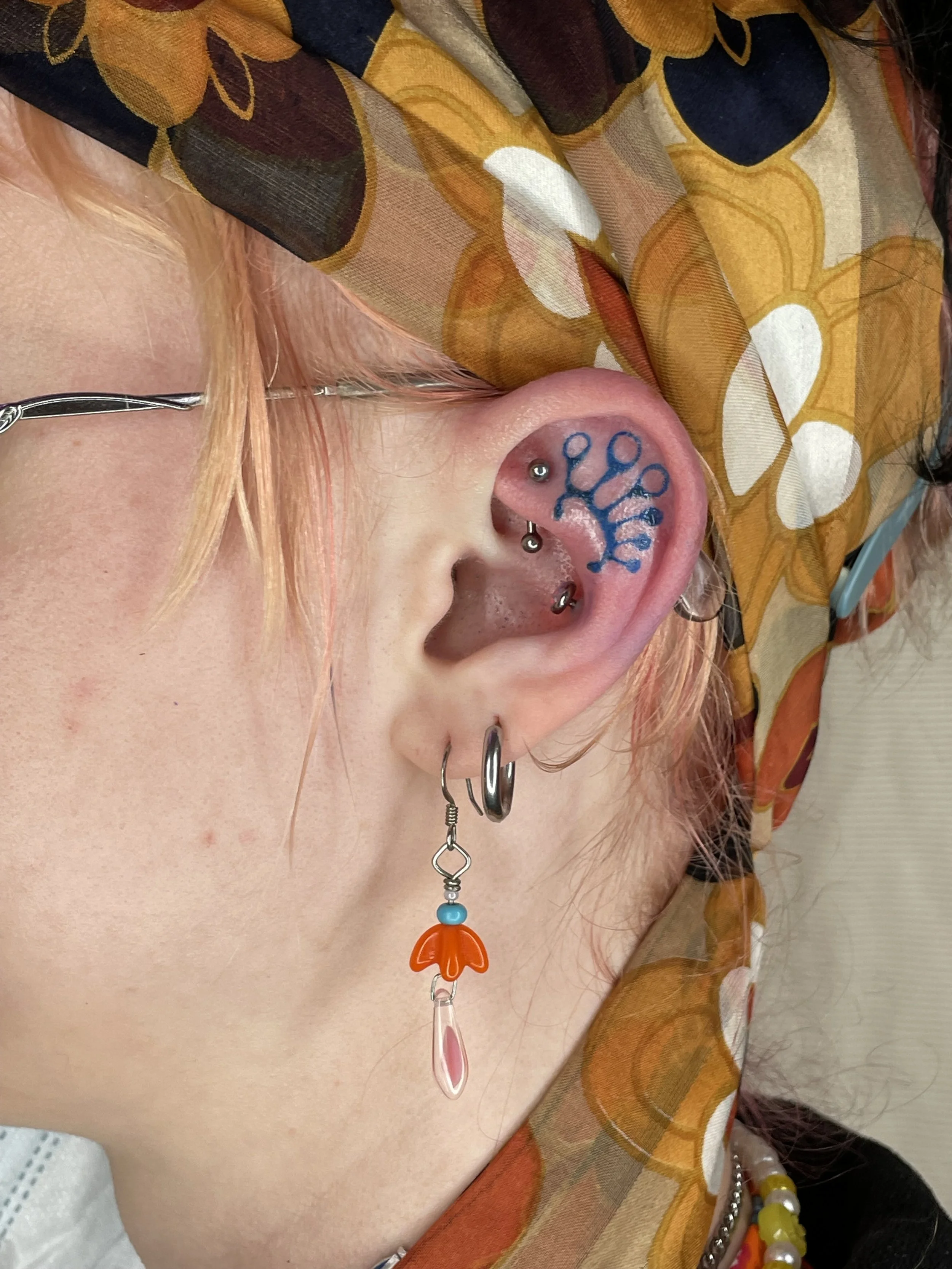 Close-up of a person with multiple facial piercings, including an industrial piercing, a nose ring, and earrings. ear tattoo, blue ink. They are wearing a colorful beaded necklace.