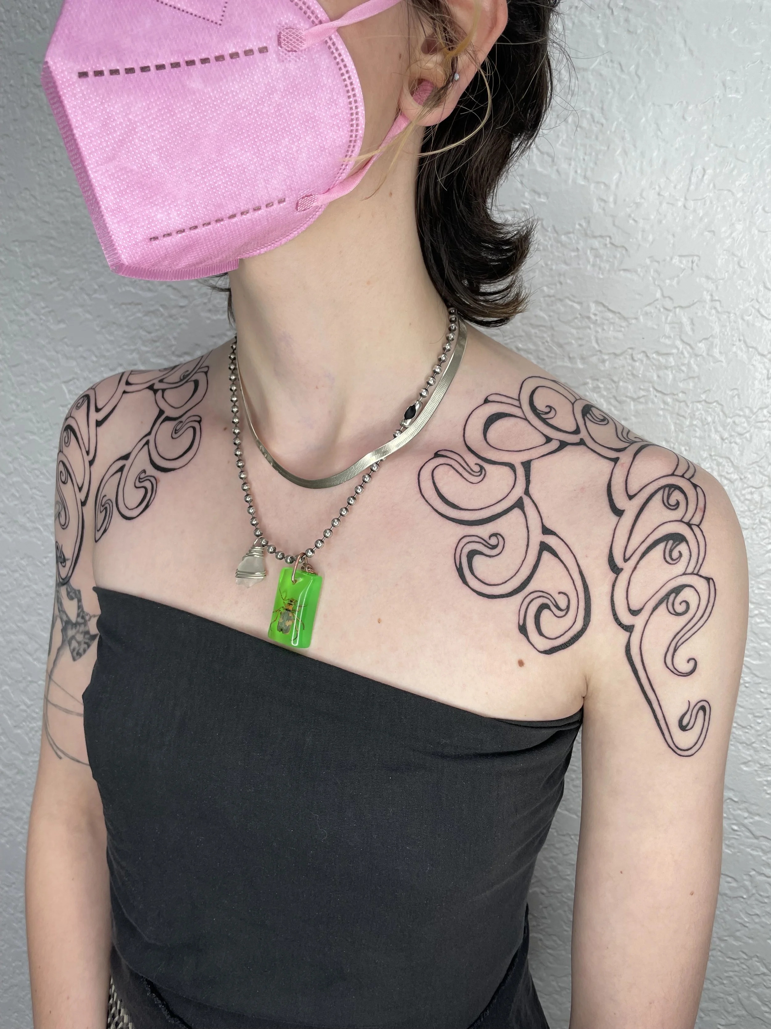 A woman wearing a pink face mask, a black strapless top, and multiple necklaces, standing against a textured gray wall. She has tattoos on her shoulder and arm, and her hair is dark and wavy.