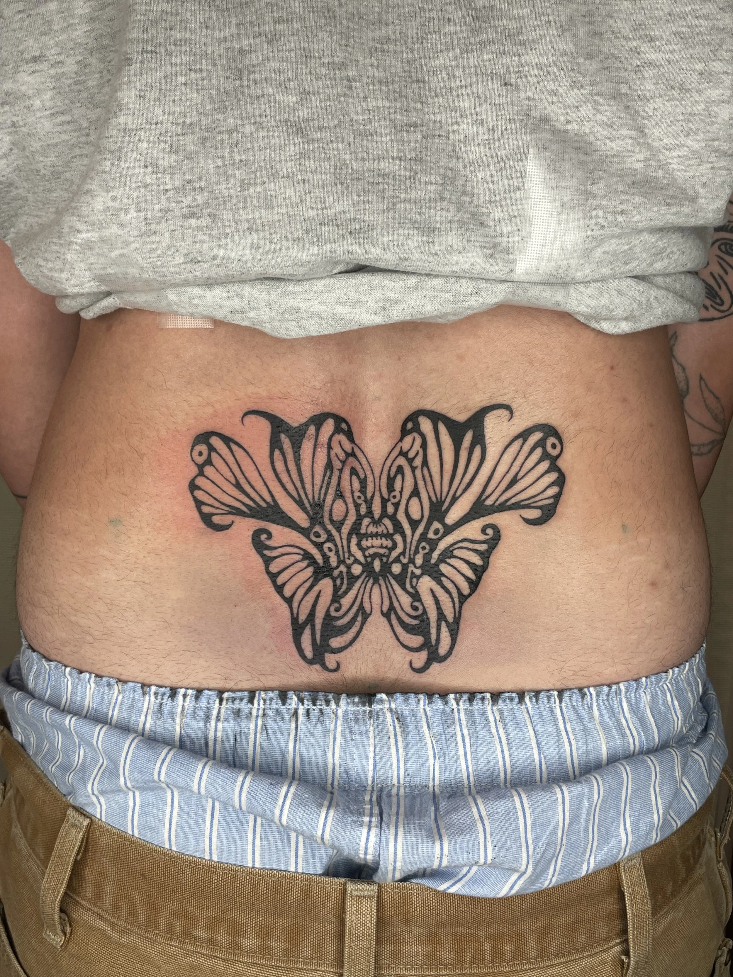 A large black butterfly tattoo on a person's lower abdomen, with a grey shirt lifted above the tattoo and blue and white striped shorts.