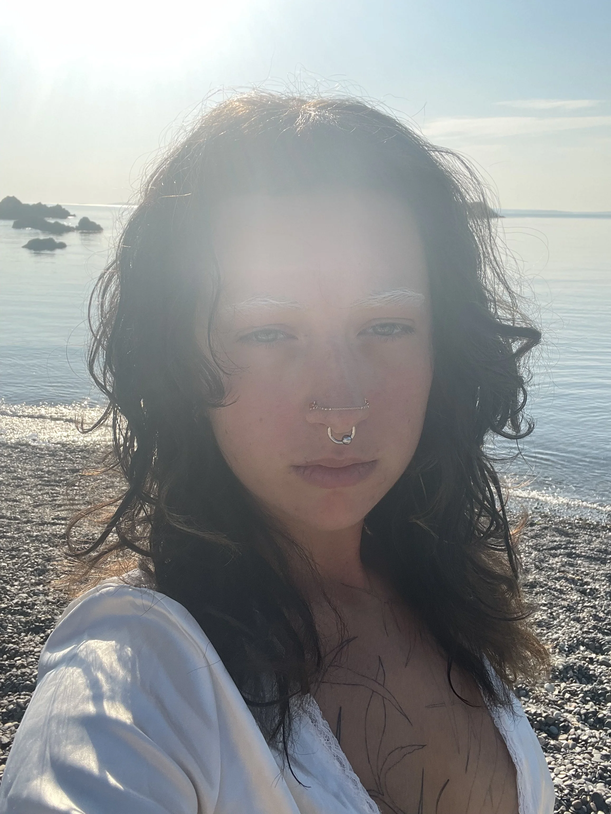 A woman with dark curly hair and septum piercings standing on a rocky beach with water and rocks in the background, sunlight reflecting on the water, wearing a white top.