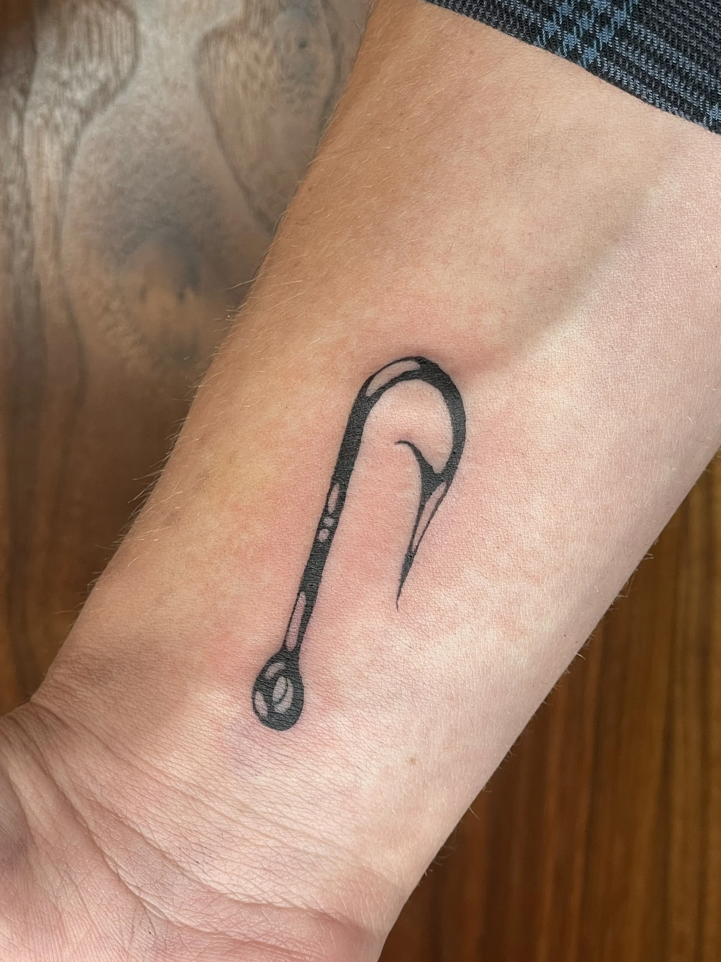 Close-up of a person's upper arm showing a black line art tattoo of a fish hook on the skin, with a background of wooden surface.