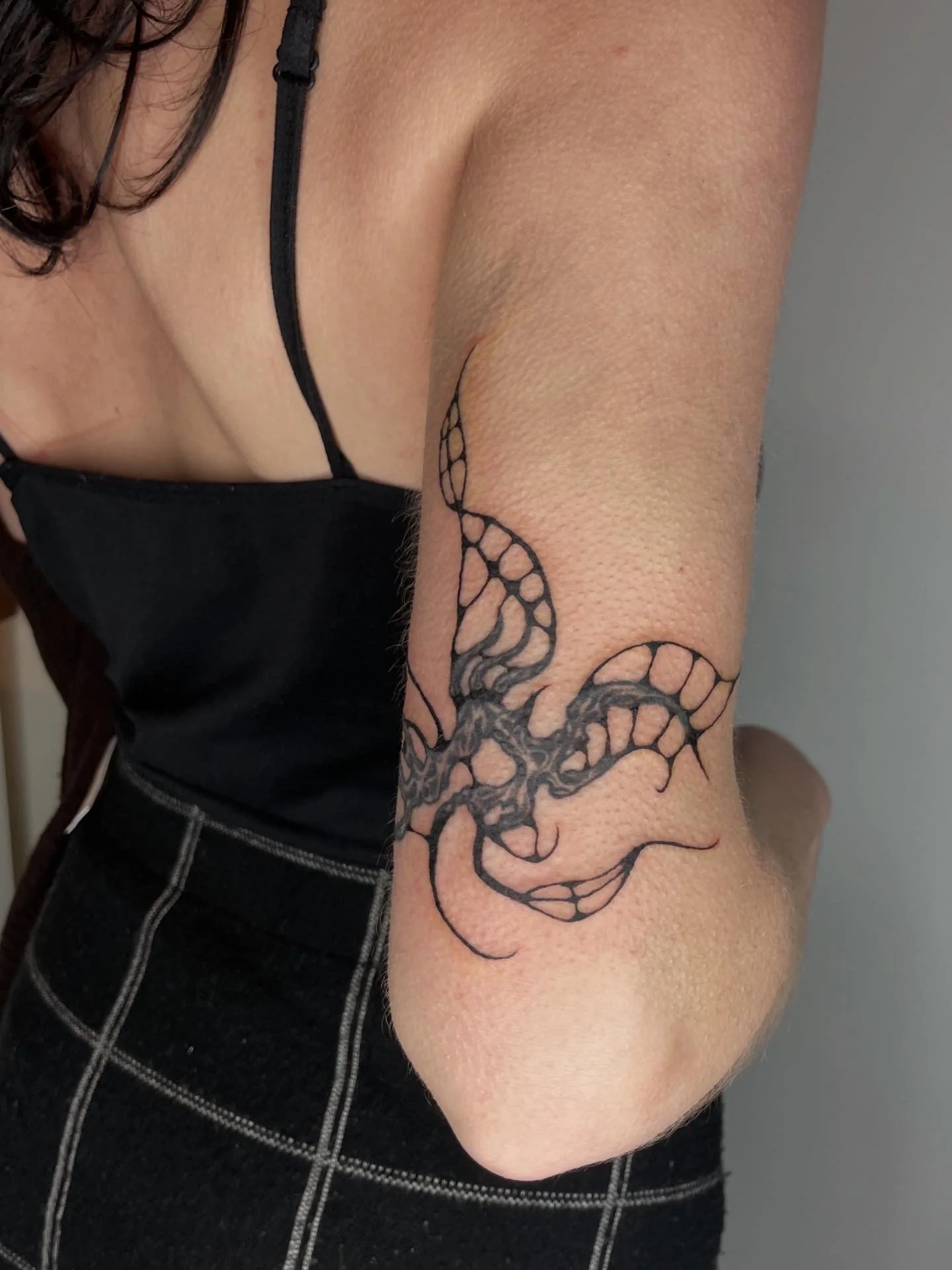 A person's arm with a black tattoo of a stylized dragon or serpent. The person is wearing a black sleeveless top with thin straps and dark pants with white stitching.