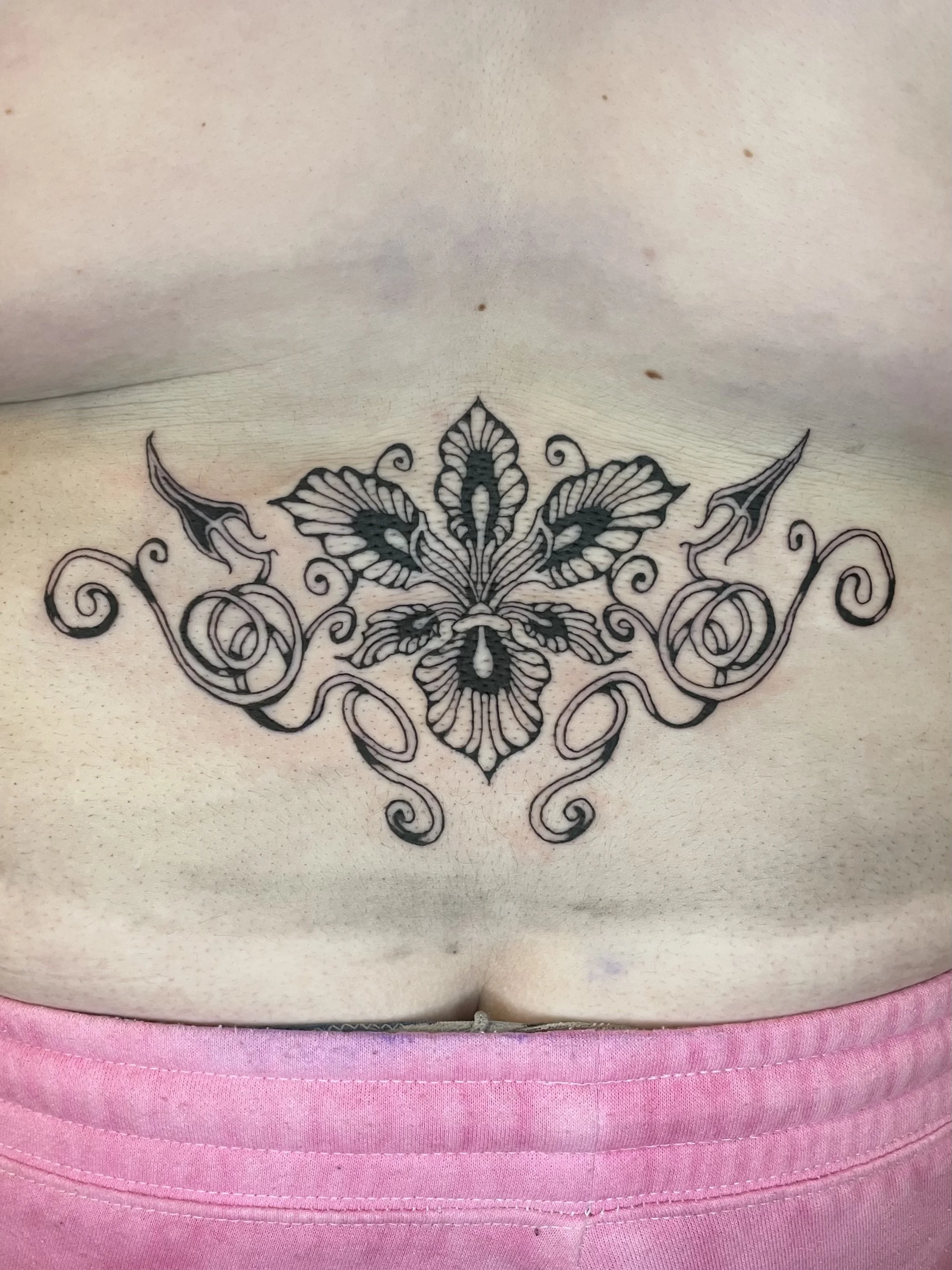 Close-up of a person's chest with a black floral tattoo just below the collarbone, wearing pink clothing.