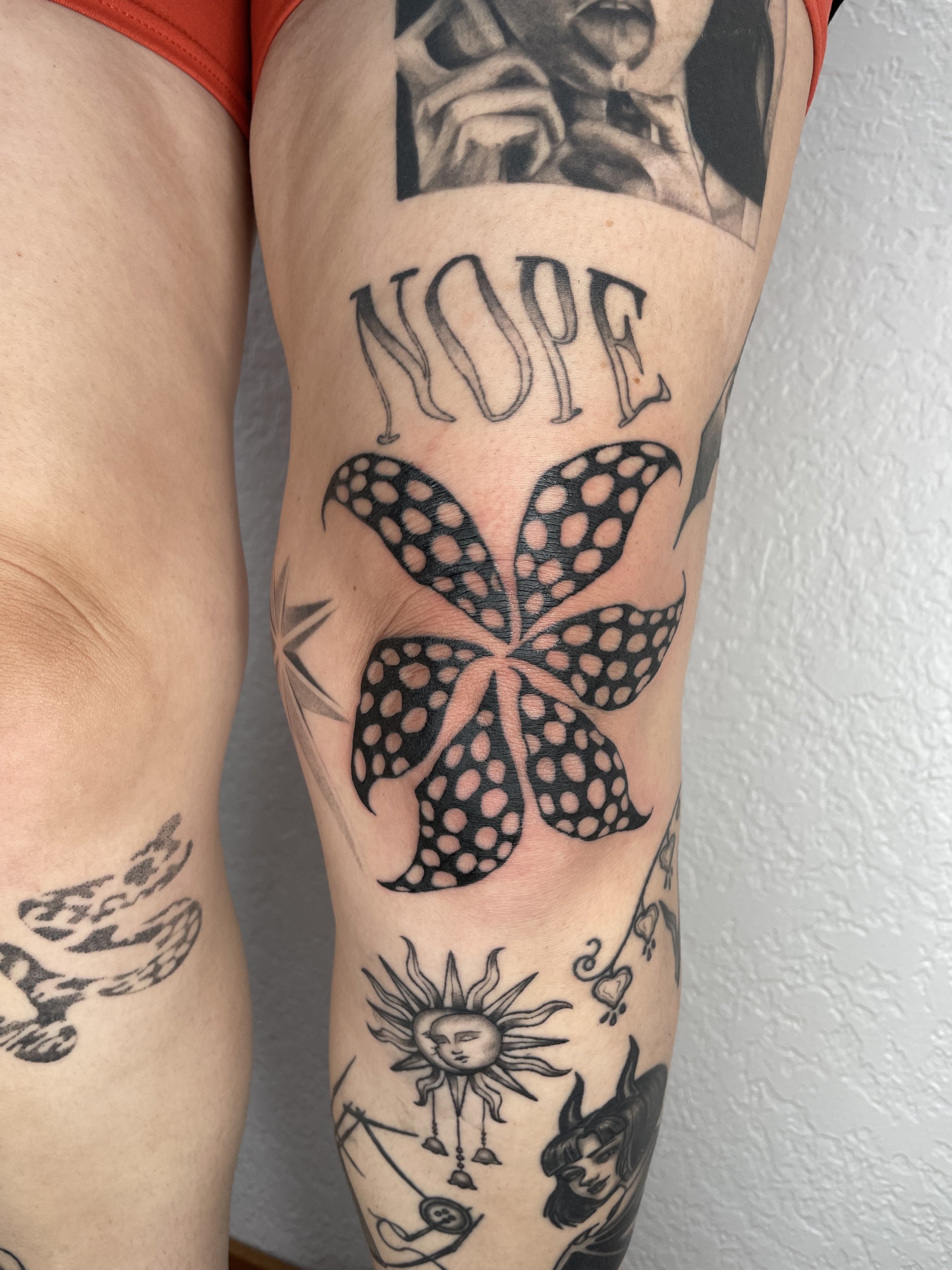 Close-up of a person's thigh with multiple tattoos, including a large butterfly with polka-dot wings, the word 'NOPE' in stylized letters, a sun with a face, and other smaller designs.