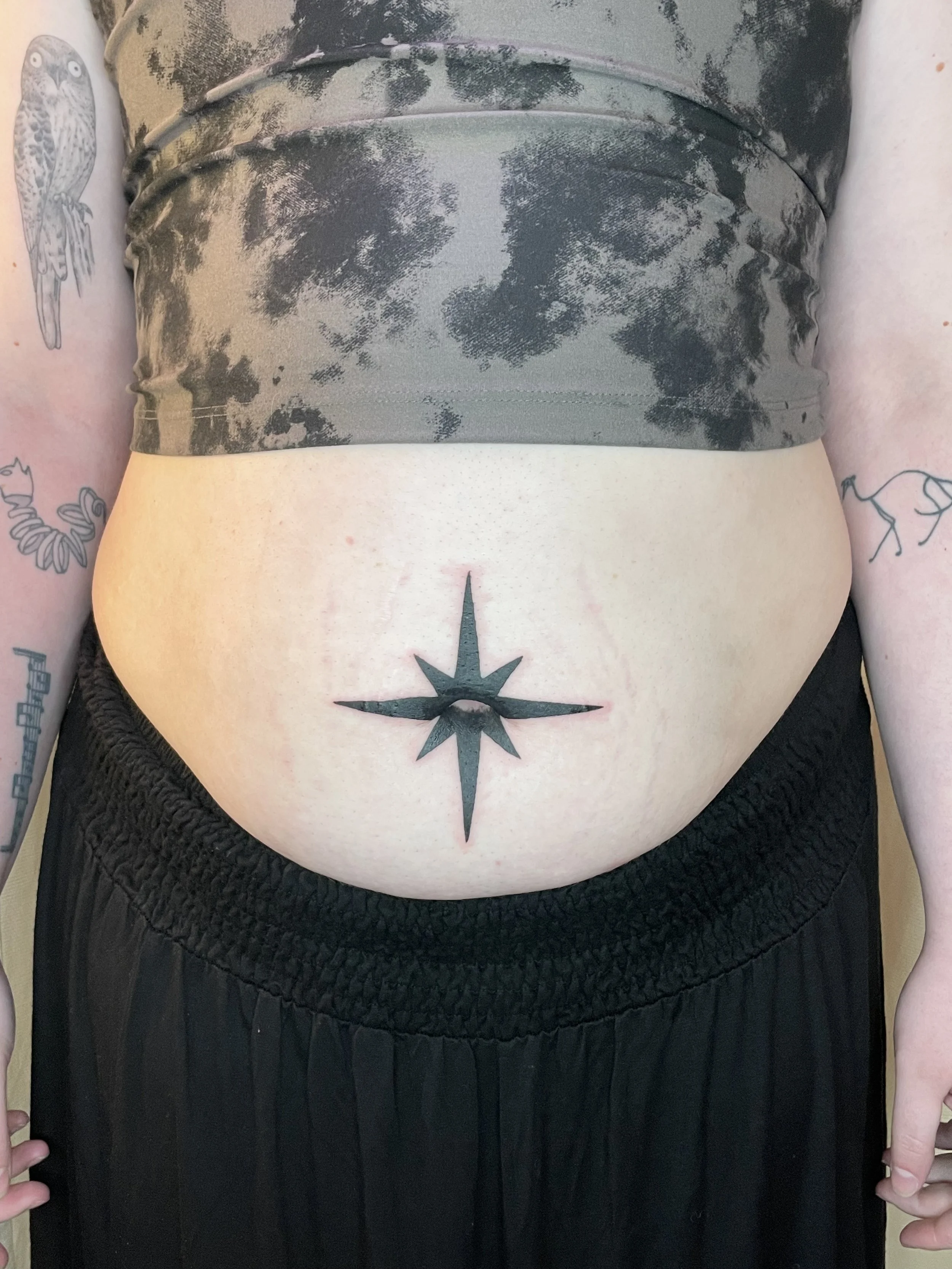 A person with tattoos on their arms and a star tattoo on their lower abdomen, wearing a tie-dye crop top and black shorts with an elastic waistband.