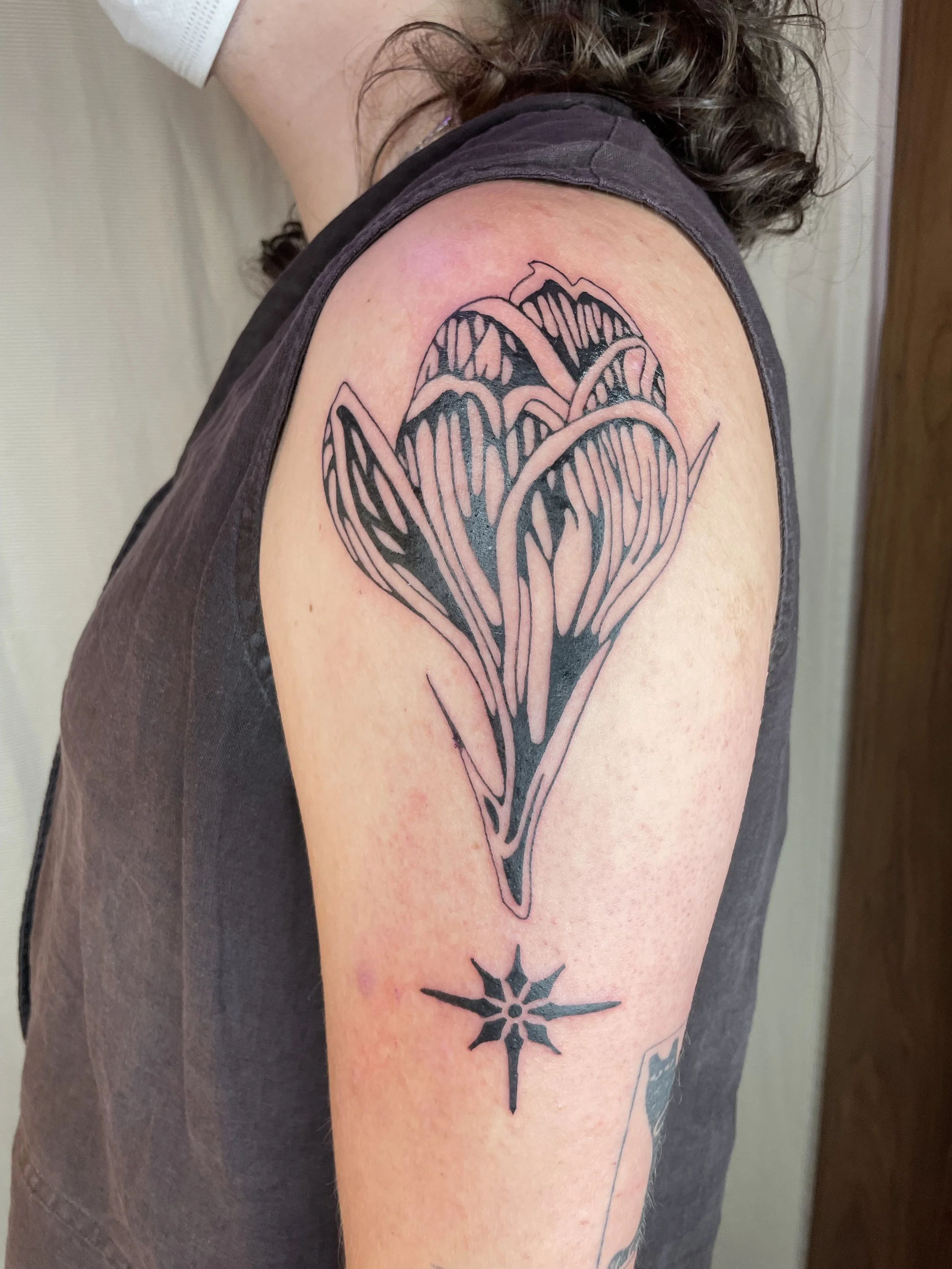 A tattoo of a flower with long, stylized petals and a star-shaped design the stem, inked in black, on a person's upper arm.