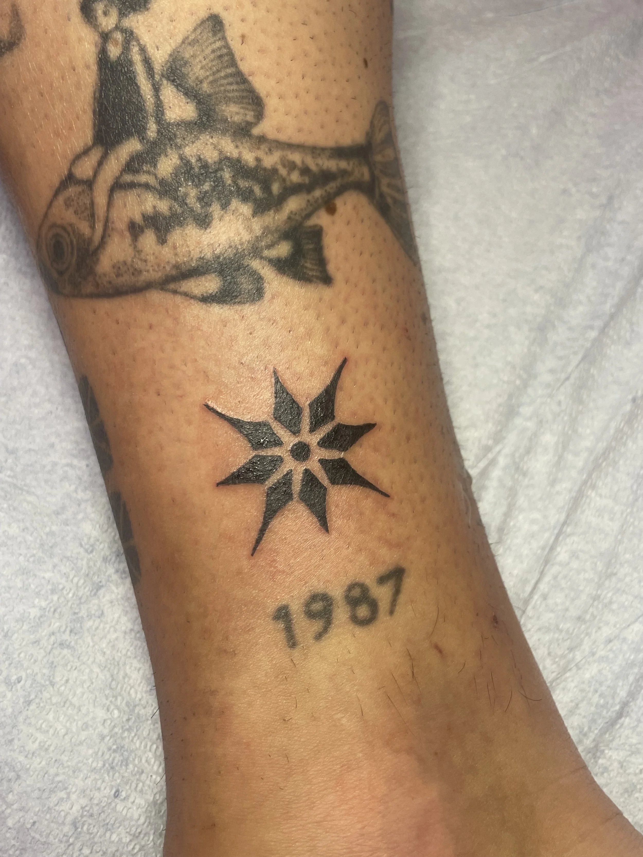 A tattoo of a fish on a person's arm, with a smaller star-shaped tattoo below it, and the numbers '1987' beneath the star.