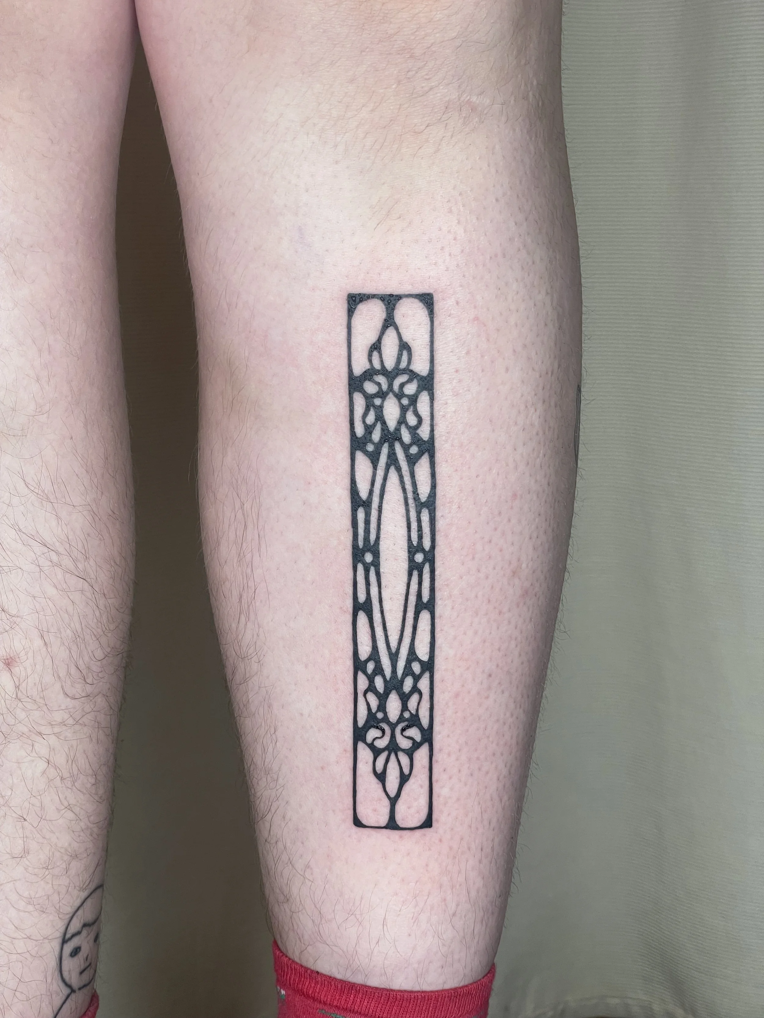 A person's thigh with a black geometric tattoo in a rectangular shape extending from the upper to lower thigh. The person is wearing red and green striped socks.
