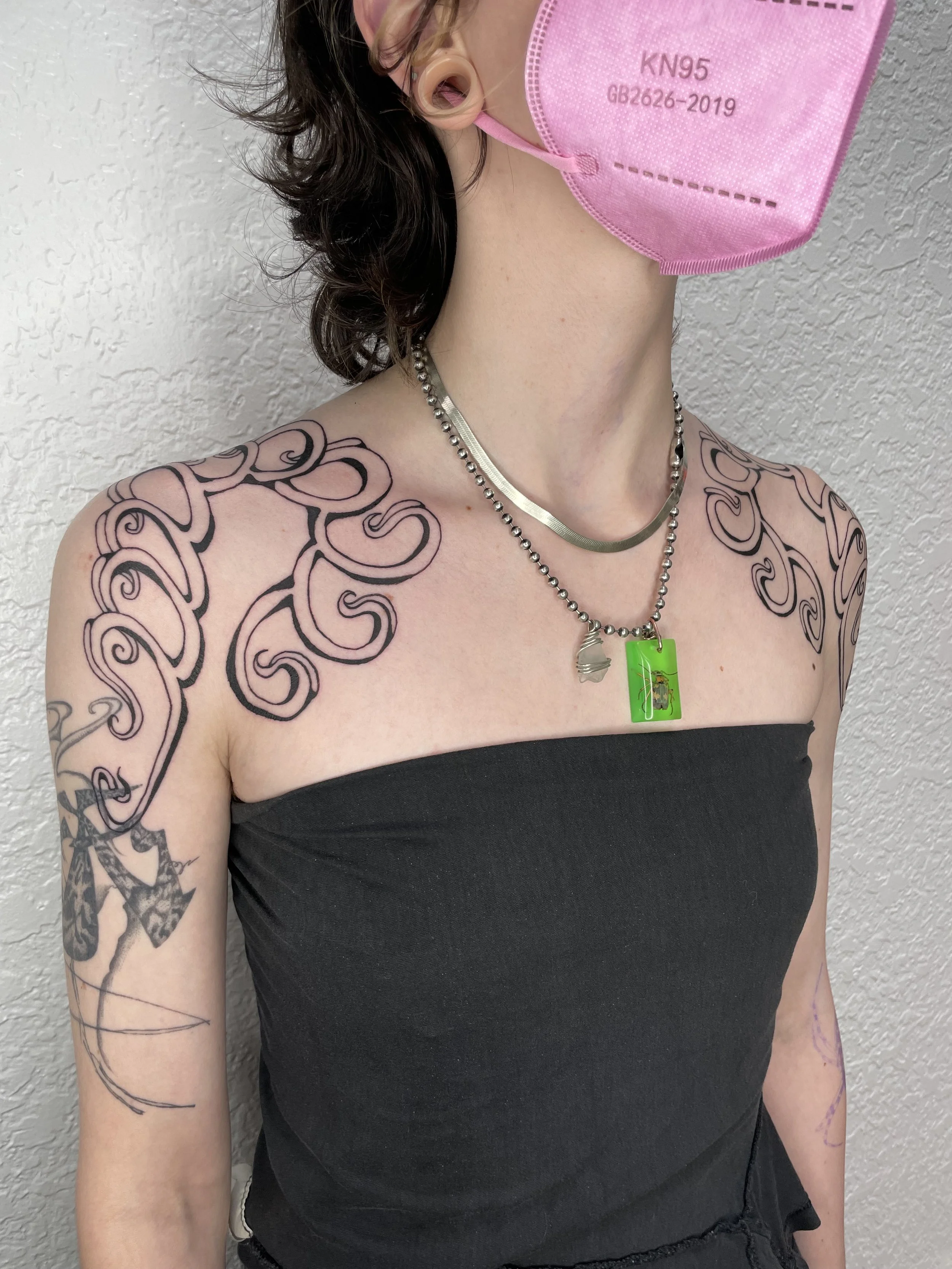 A person with shoulder-length, curly dark hair wearing a pink KN95 face mask, silver necklaces, and a black strapless top, standing against a textured white wall with tattooed arms.