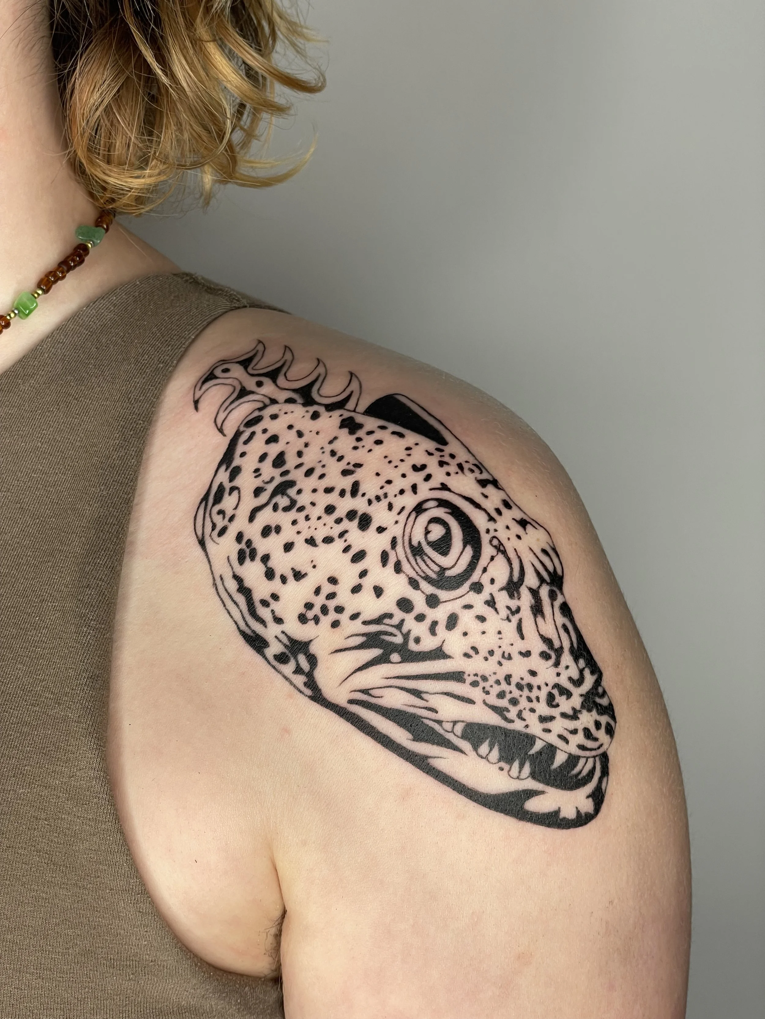 Close-up of a person's upper arm with a black ink tattoo of a lingcod with detailed patterns, dressed in a necklace with brown and green beads, and wearing a brown sleeveless top.