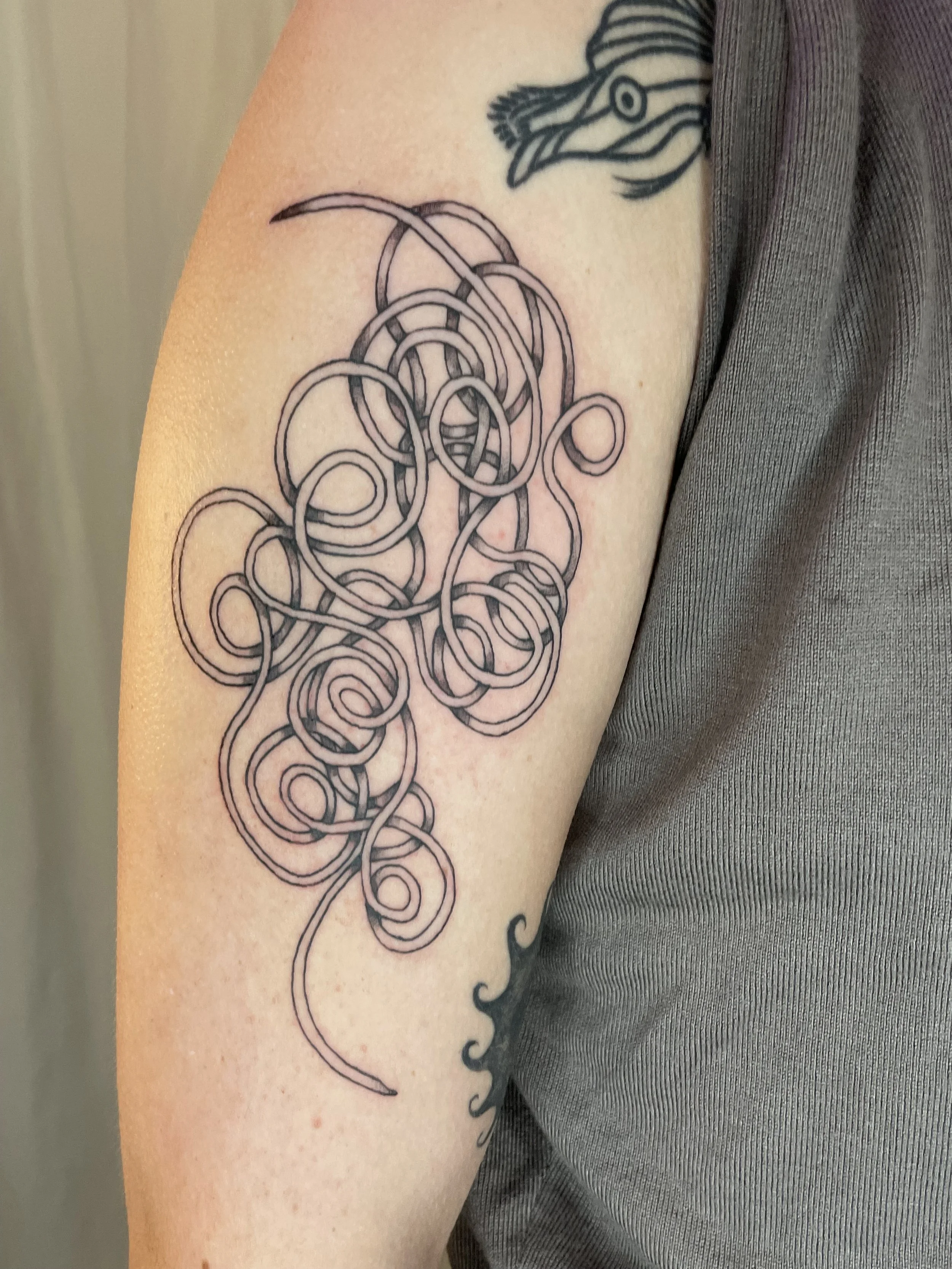 A tattoo at the top of a person's arm, abstract black line art of a mass of intertwined, looping lines extending downward.