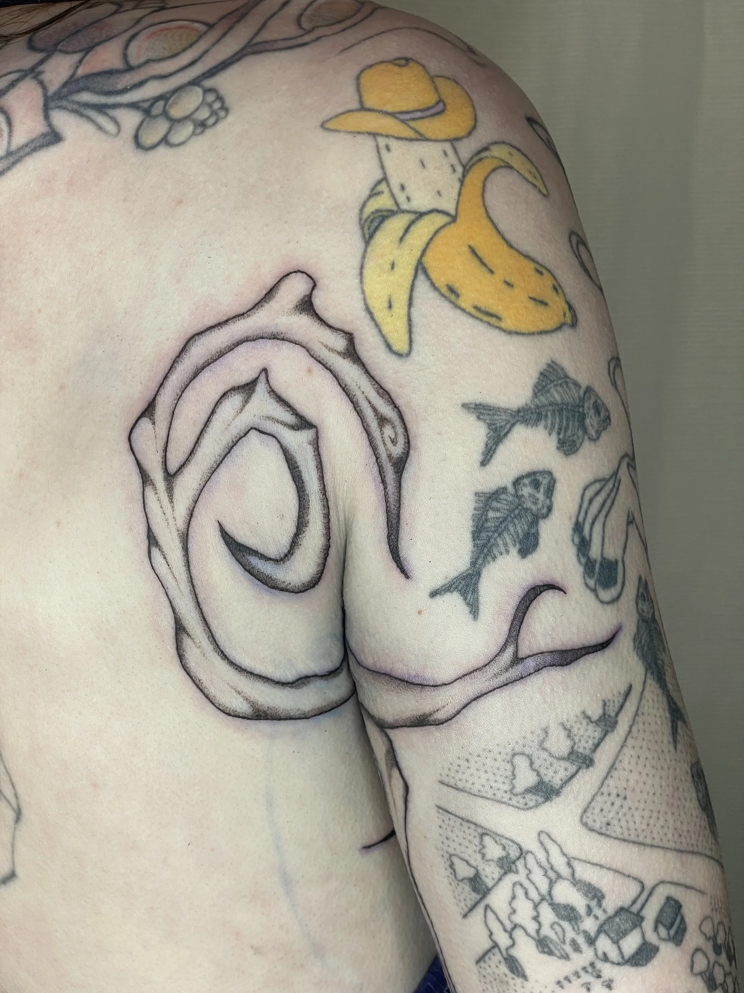 A person's shoulder and upper arm with various tattoos, including a yellow cowboy hat and a banana, along with other designs and characters.