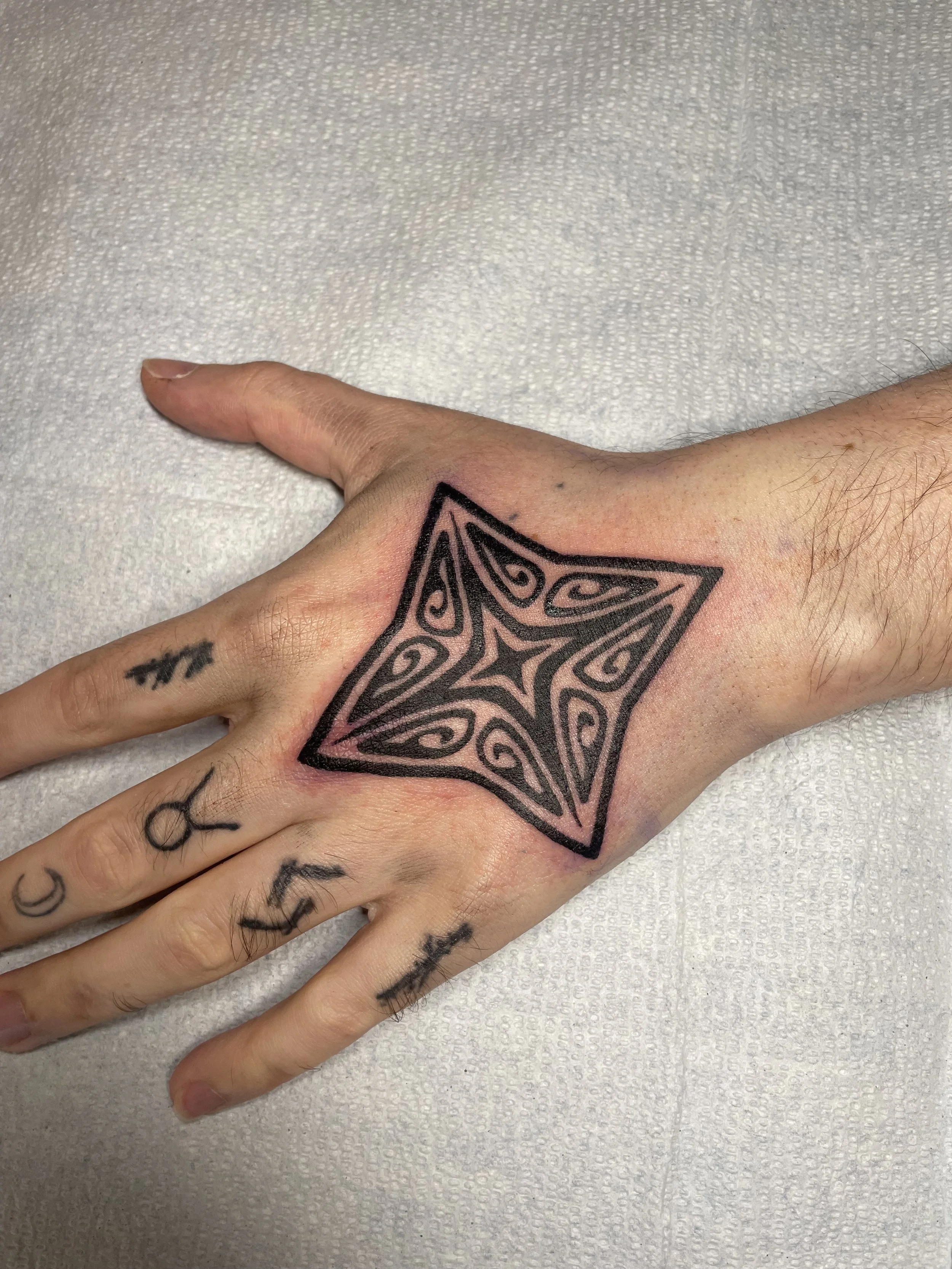 A hand with tattoos resting on a white textured surface, featuring a large black tribal star tattoo on the back of the hand and smaller tattoos on the fingers.