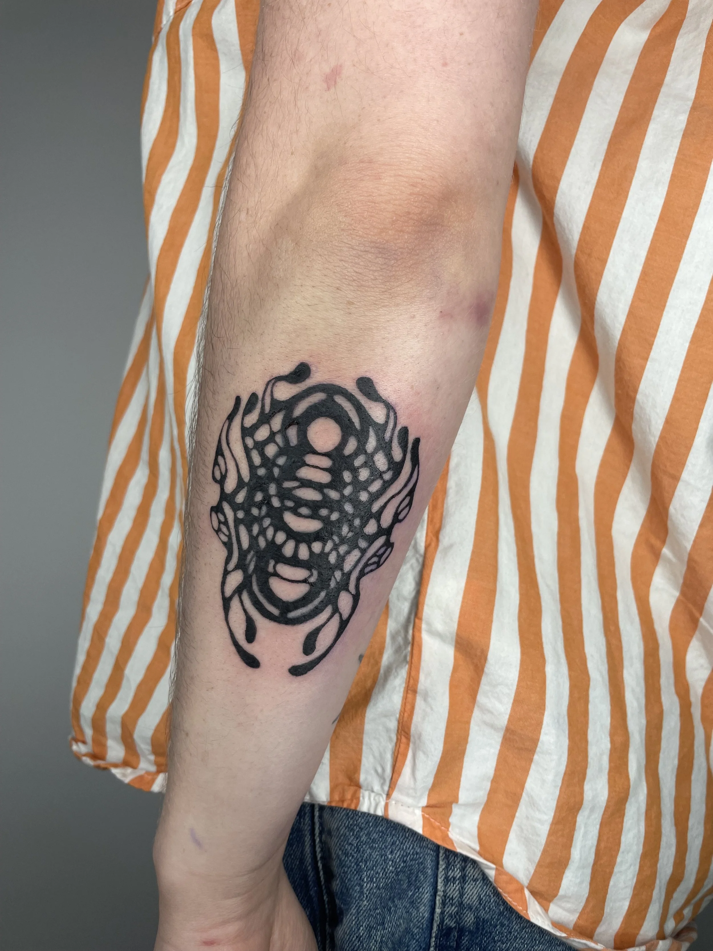 Close-up of a person's forearm with a black abstract tattoo resembling a tribal or artistic design. The person is wearing a short-sleeved shirt with orange and white vertical stripes and blue jeans.