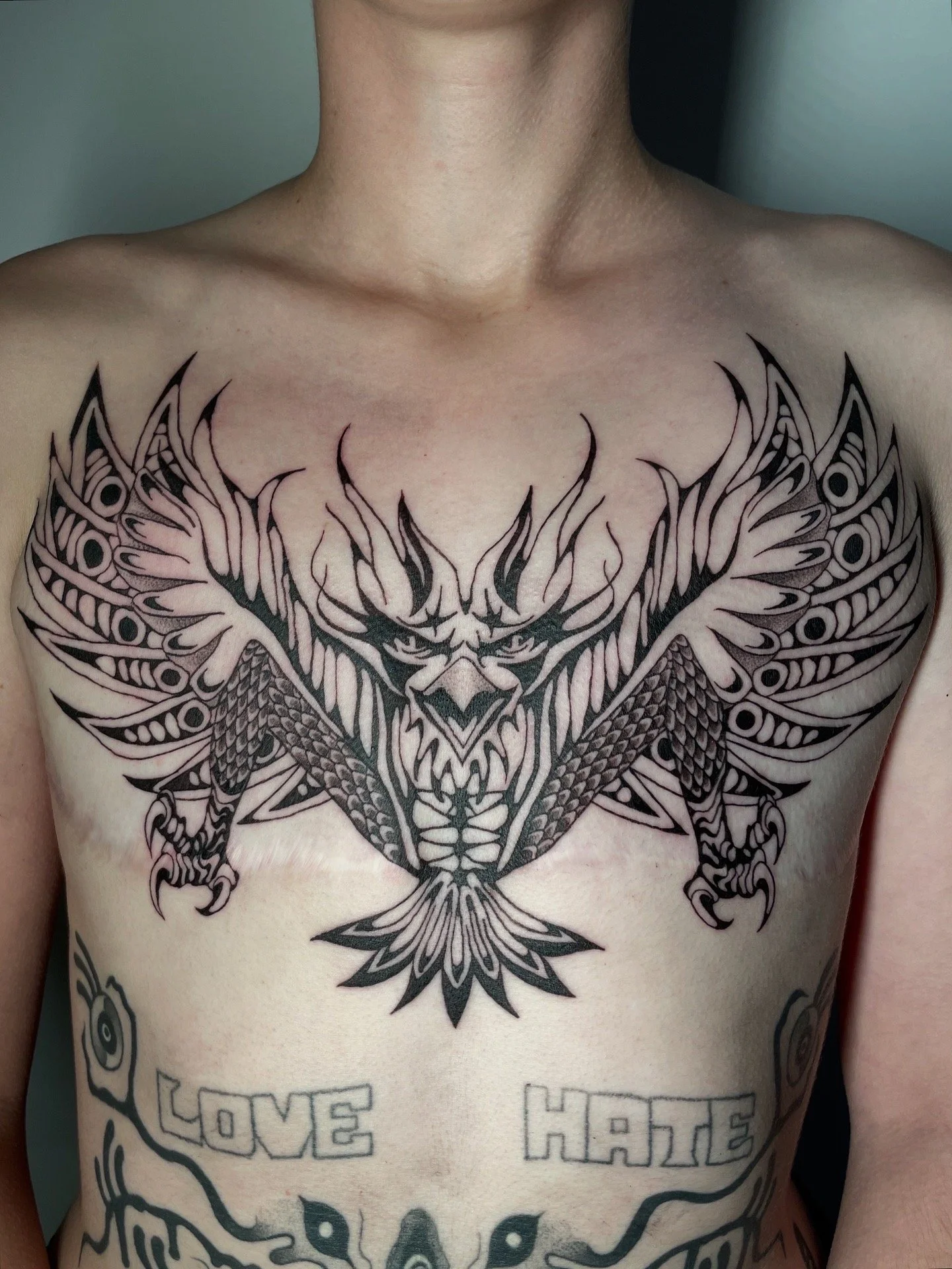 Black tribal-style tattoo of a butterfly with detailed wings and a face in the center, on a person's chest with words 'LOVE' and 'HATE' below.
