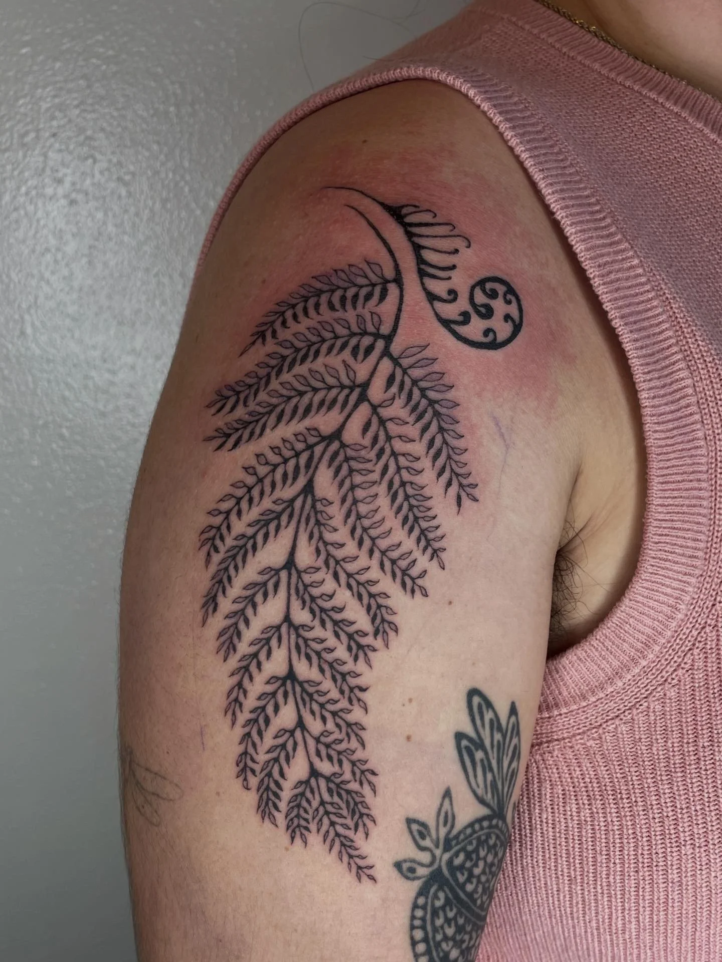 Tattoo of a large, intricate fern leaf design with a smaller, circular decorative element near the top on the upper arm of a person wearing a pink sleeveless shirt.