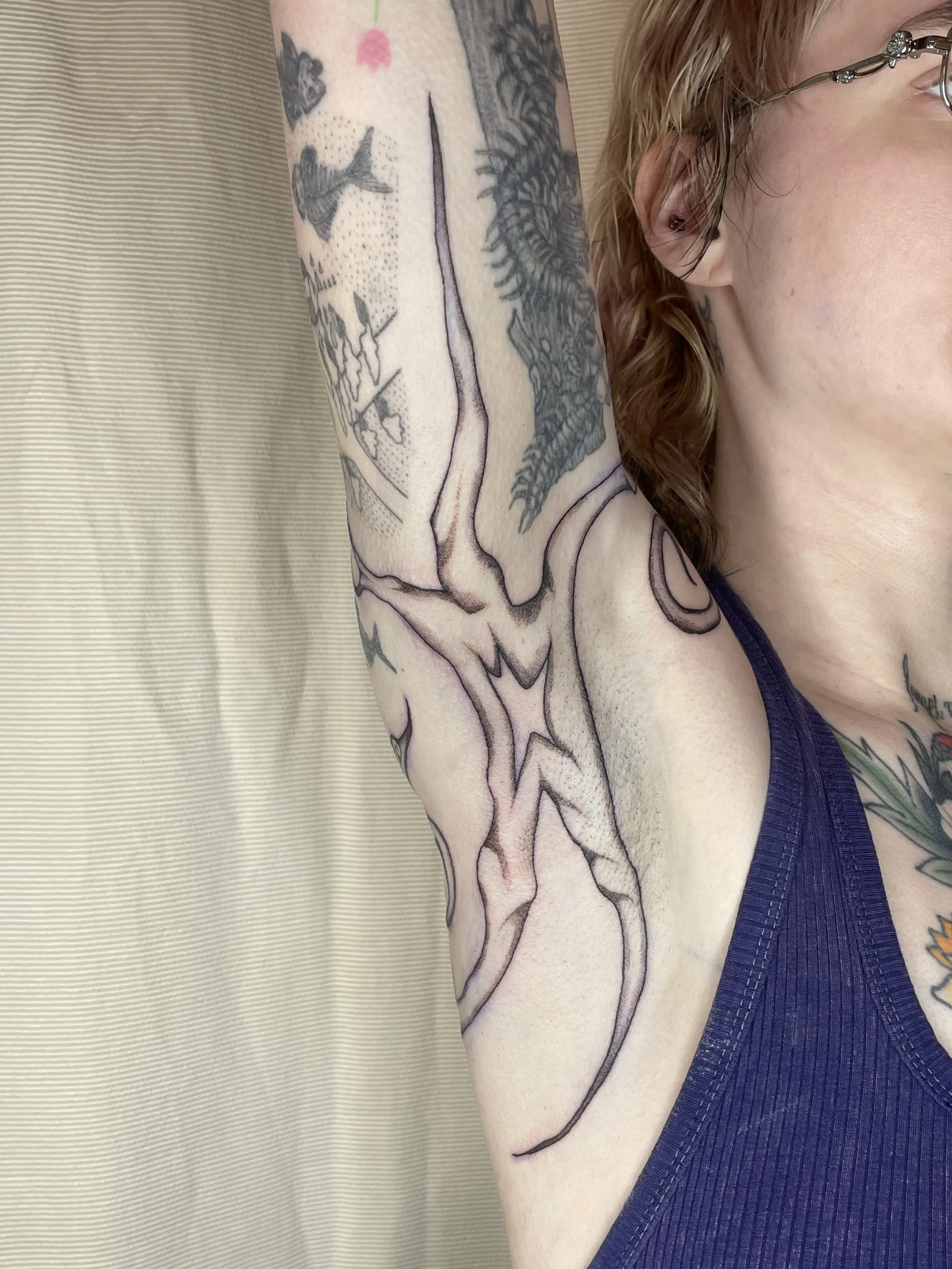 Close-up of a person's arm with detailed black and gray tattoos, including a bird, abstract designs, and patterns, wearing a purple sleeveless top, against a neutral background.