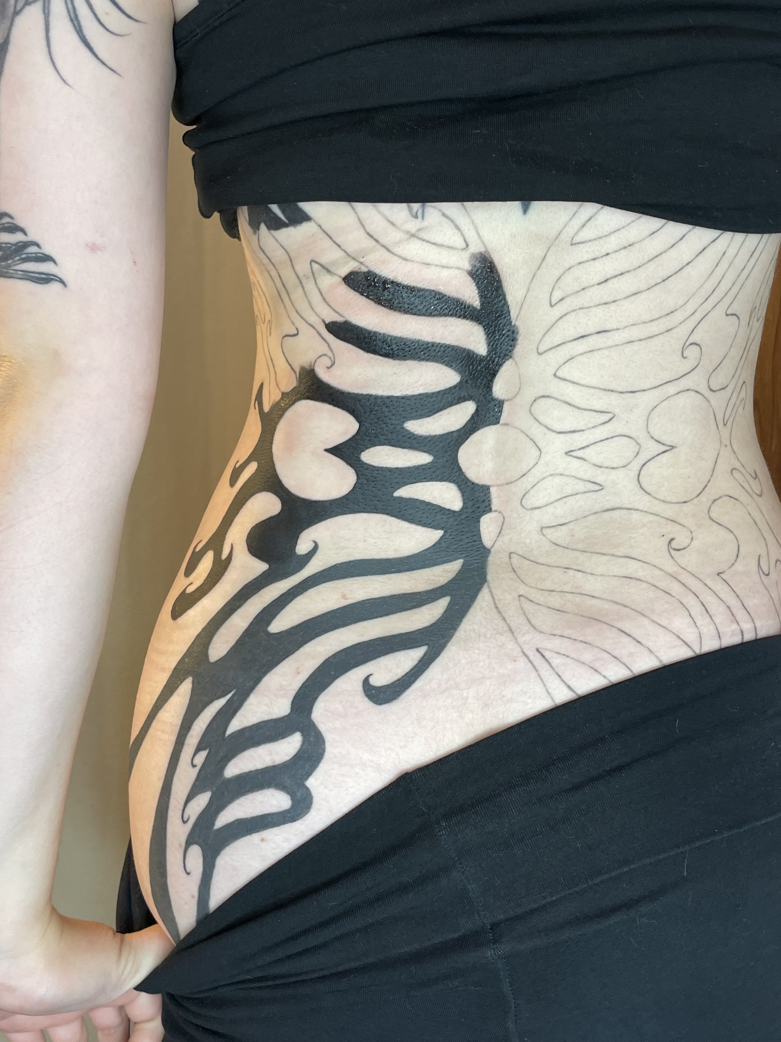 Close-up of a person's side torso showing large black and white abstract tattoo covering the side of the ribcage, with part of the person's arm and black clothing visible.