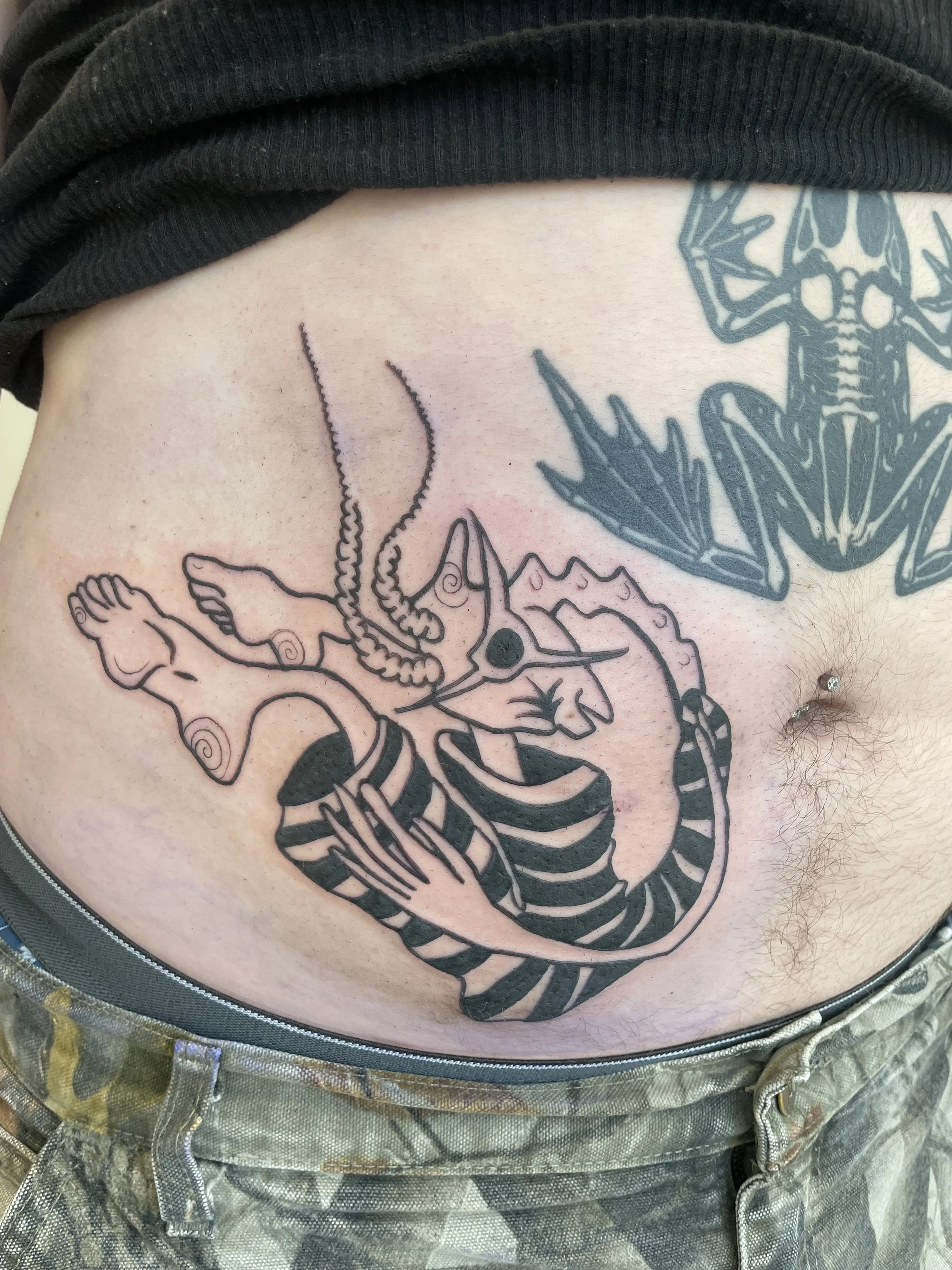 Tattoo of a skull with octopus tentacles on person's abdomen, partially covered by black clothing and camouflage shorts.
