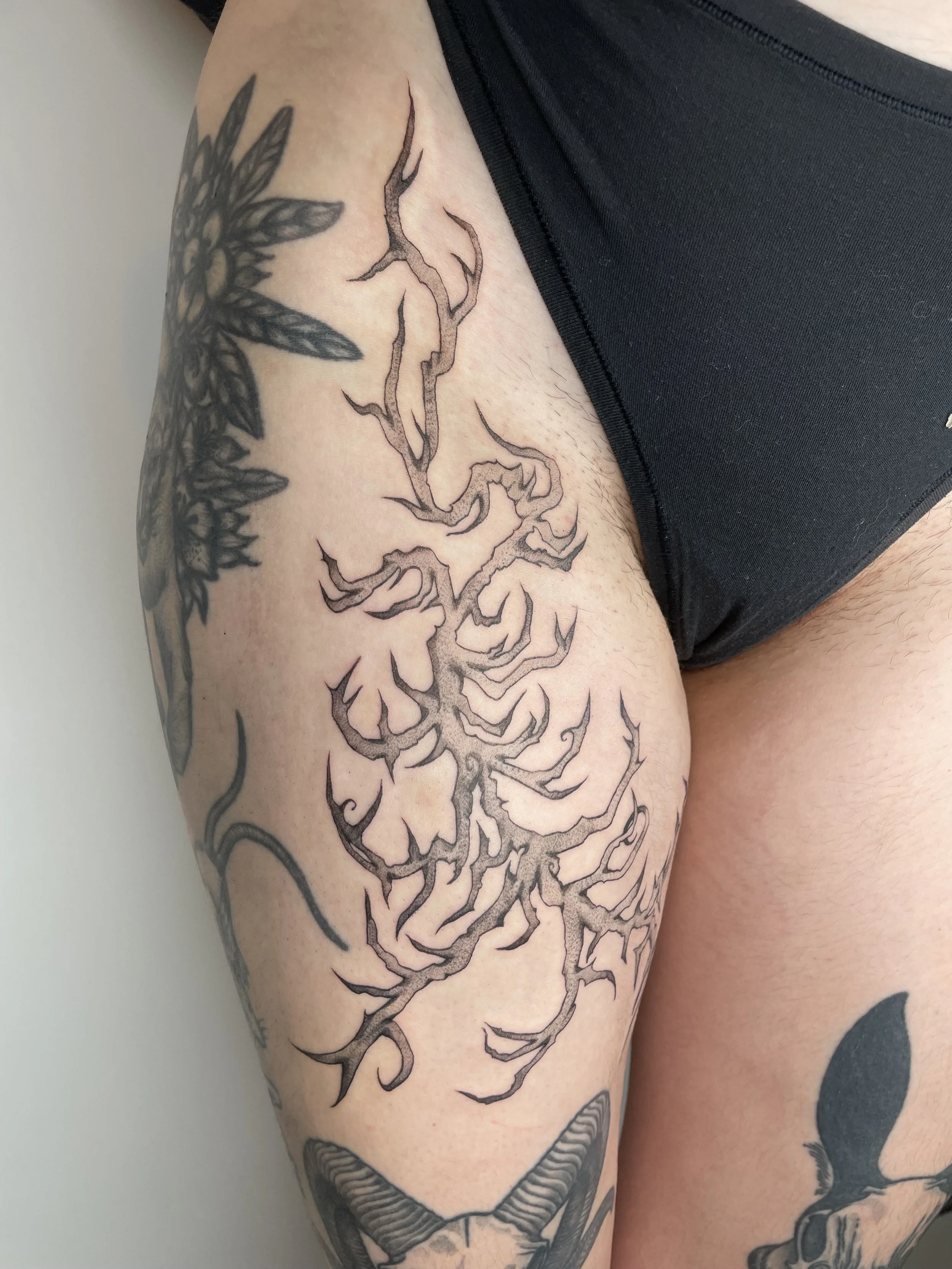 A person's upper arm features a detailed black and grey tattoo of a tree with numerous branches. Additional tattoos are visible, including a large floral design and other elements.