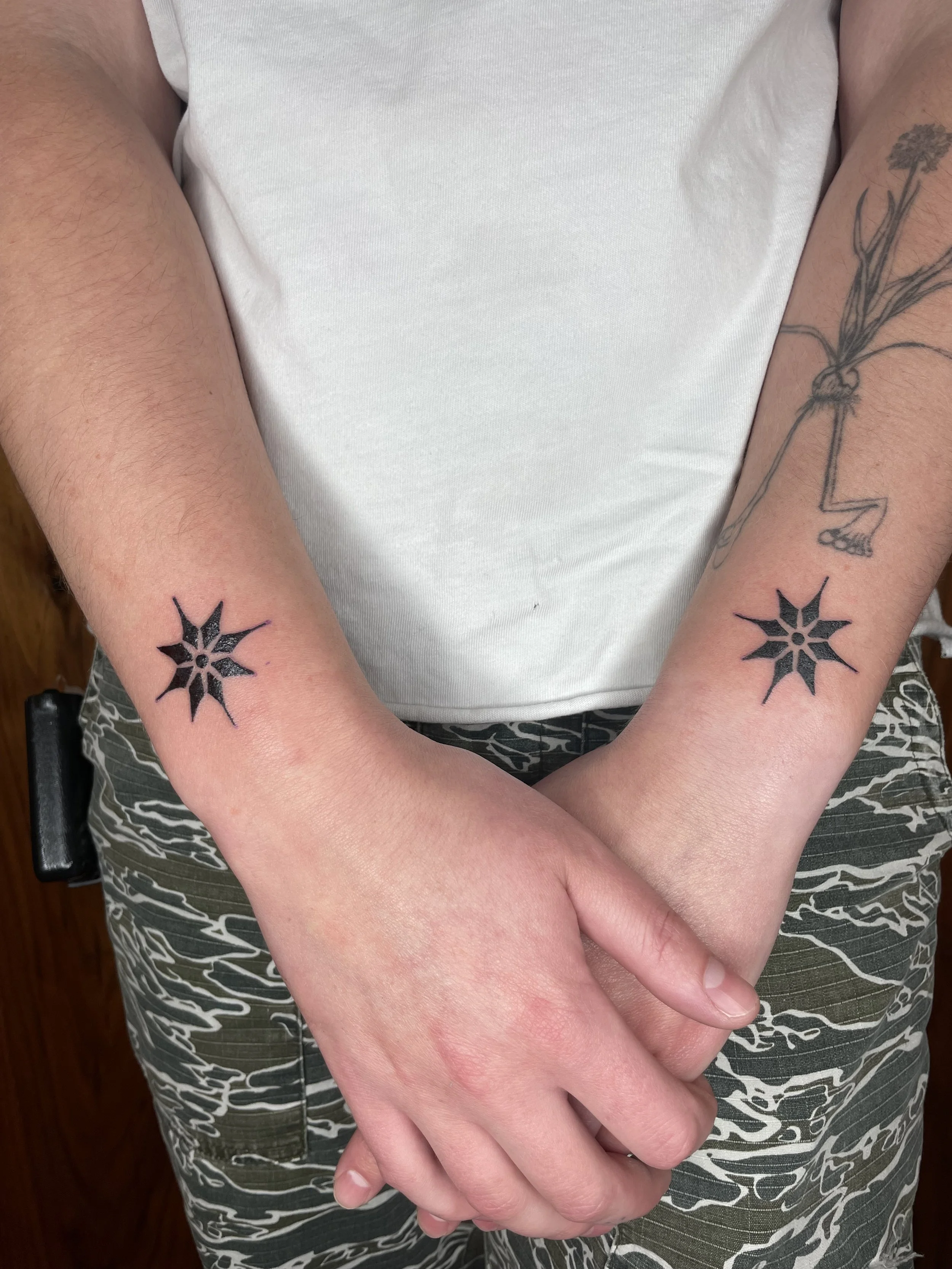 Close-up of a person's arms and hands crossed over each other, wearing a white t-shirt and patterned pants, showing tattoos on their arms, including star designs on the wrists and a floral design on the left forearm.