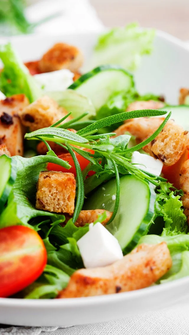 Close-up of a fresh salad with cucumbers, cherry tomatoes, croutons, pieces of grilled chicken, and fresh herbs.