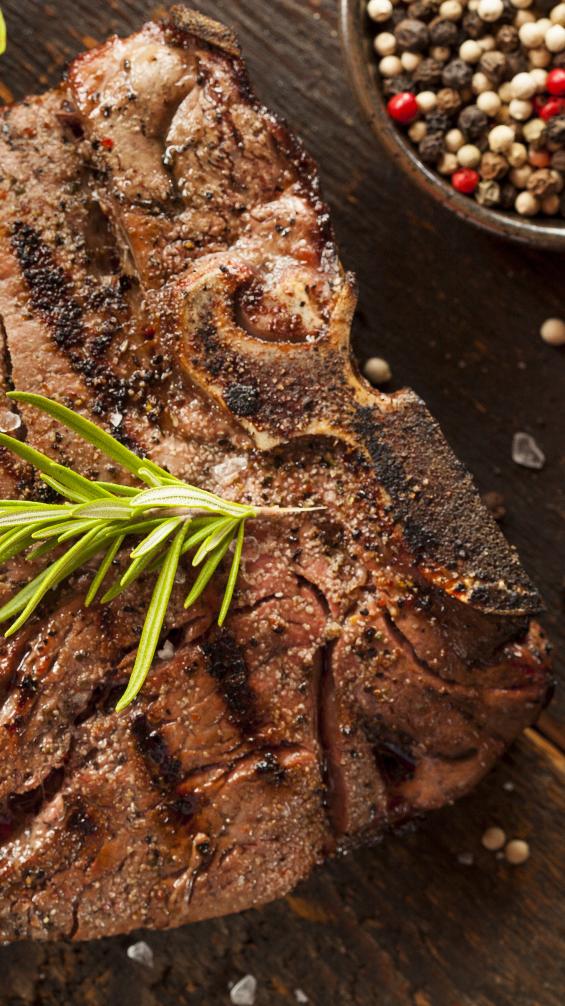 Close-up of a cooked beef rib seasoned with spices, garnished with a sprig of rosemary, and resting on a wooden surface, with a bowl of mixed peppercorns in the background.