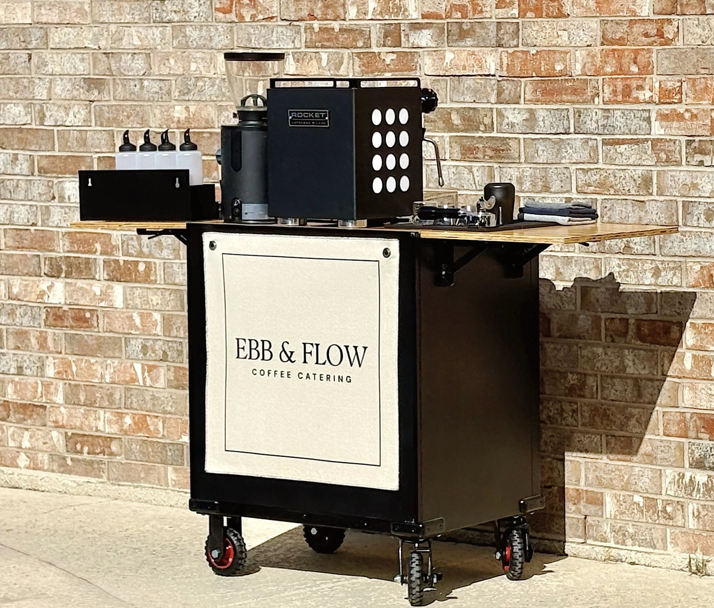 A mobile coffee cart with an espresso machine, espresso grinder, and barista tools. On the front of the cart is a banner that reads, "EBB & FLOW Coffee Catering."