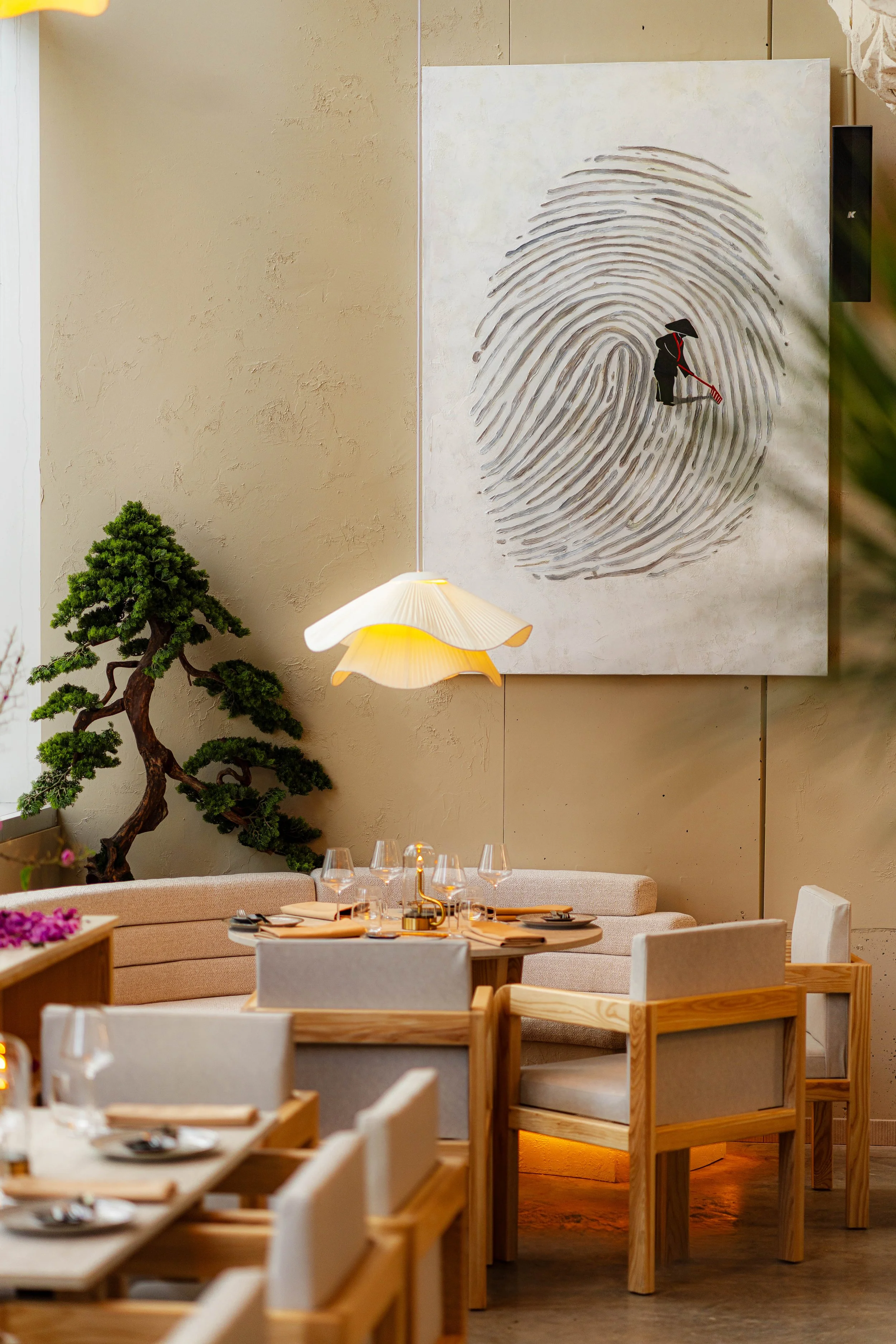 A cozy restaurant interior with a beige upholstered booth, wooden chairs, a round table set with plates, glasses, and utensils, a lamp hanging from the ceiling, an art piece featuring a fingerprint with a small figure using a rake, a bonsai tree, and a wall with textured paint.