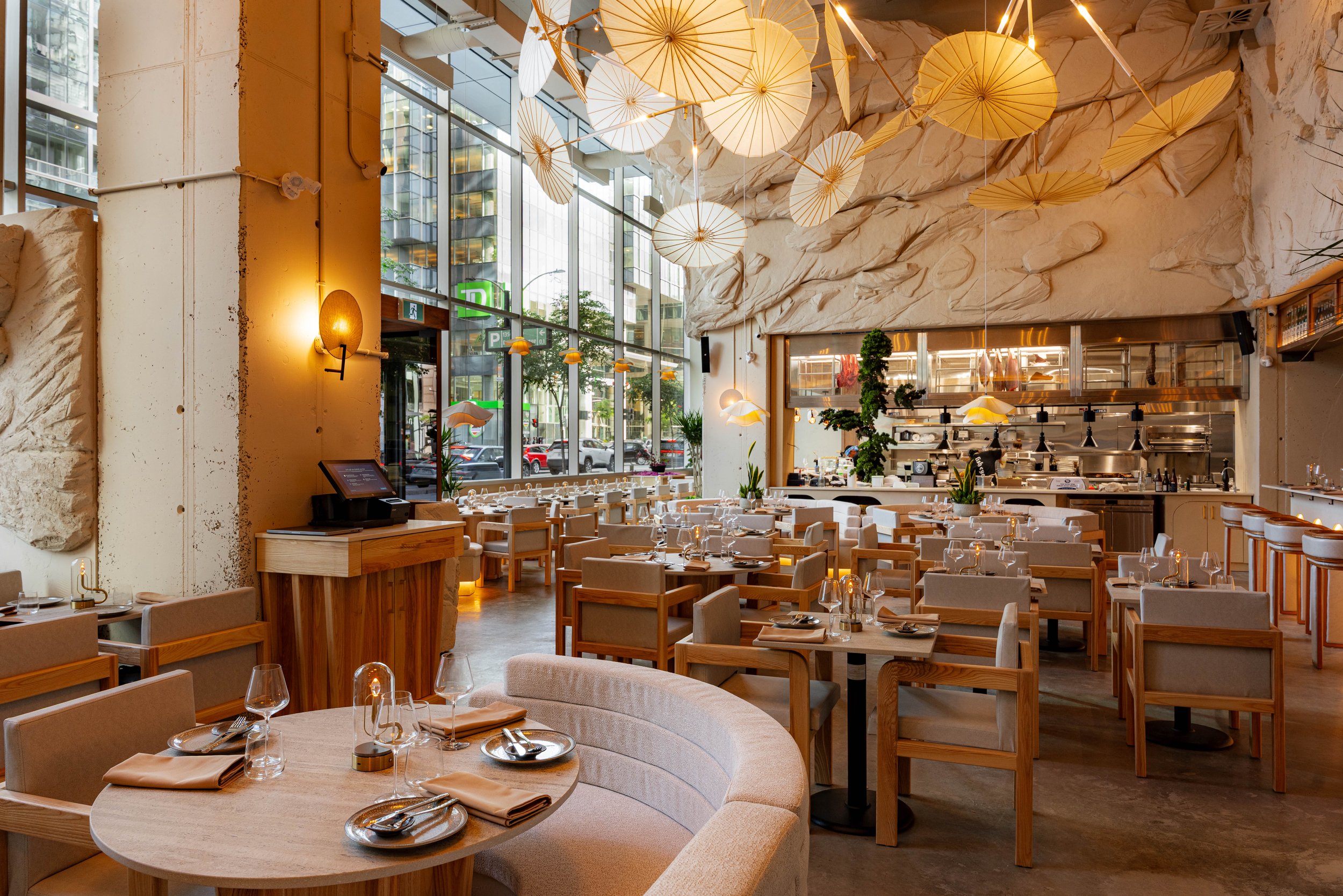 Interior of a modern restaurant with high ceilings, large windows, and decorative paper umbrellas hanging from the ceiling. Dining tables are set with glasses, napkins, and utensils, and the space features a mix of wooden and upholstered furniture.