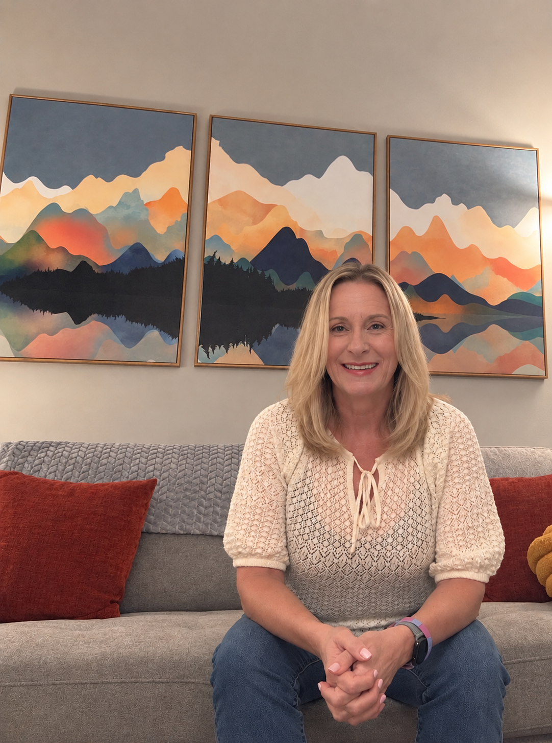A smiling woman with blonde hair sitting on a gray sofa with red and orange pillows, in front of a triptych painting of colorful mountain landscape with lakes.