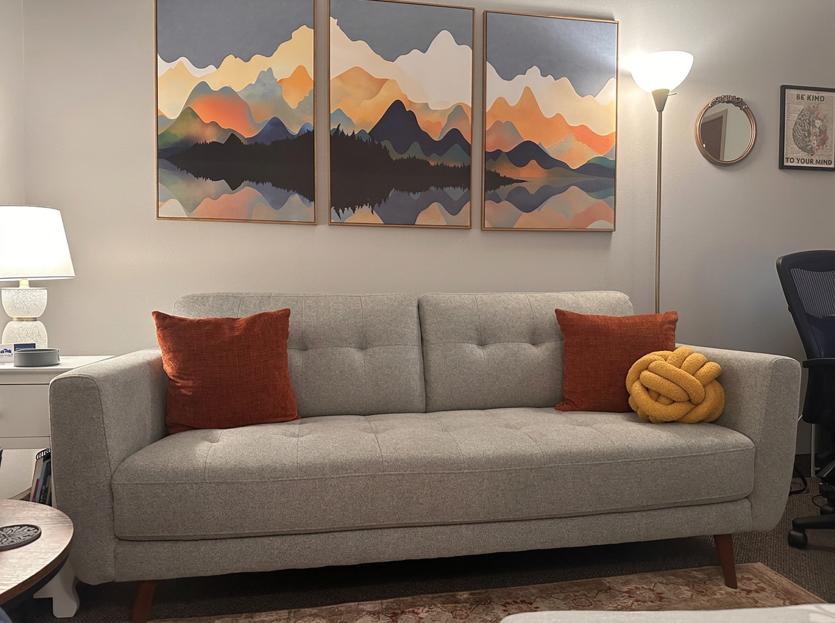 Living room with a light gray sofa decorated with two rust-colored pillows and a yellow knot pillow, a multi-panel mountain landscape art above the sofa, a white side table with a lamp and small decor items, a floor lamp, a round mirror, a framed inspirational quote, and a black office chair on the right.
