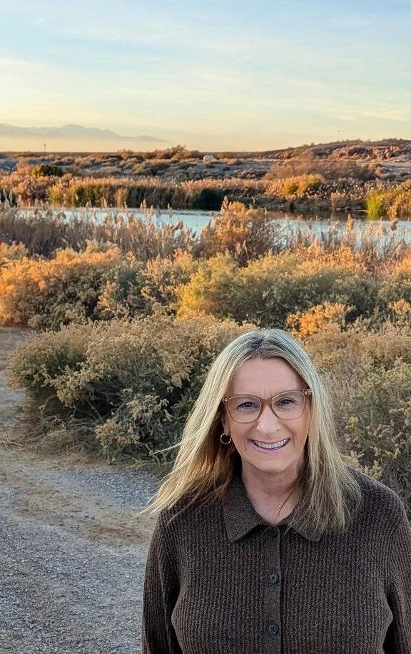 A woman with glasses and blonde hair smiling outdoors during sunset, standing on a dirt path with shrubs and a body of water in the background.