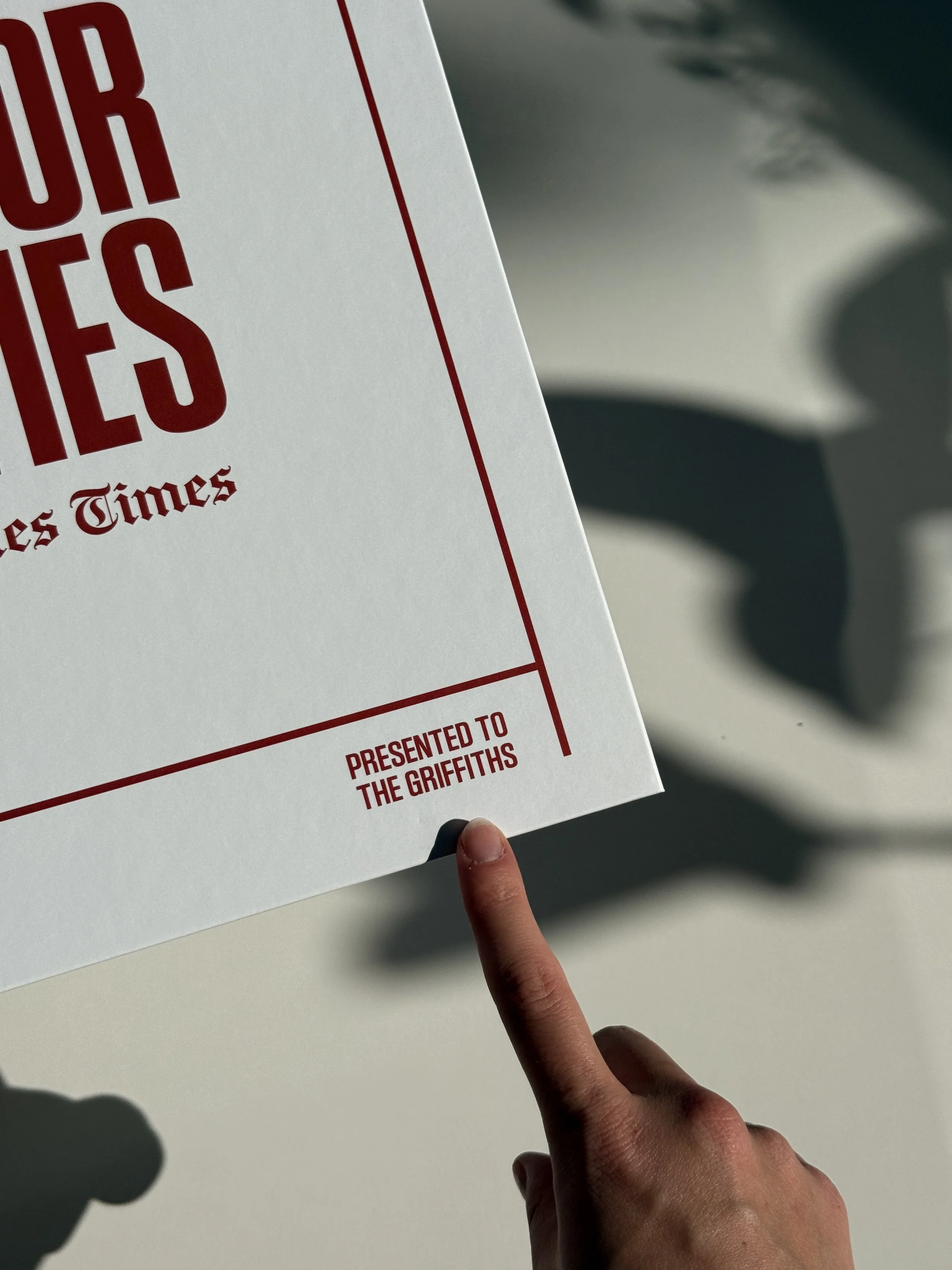 Close-up of a hand with fingers forming a peace sign, pointing at a white piece of paper or cardboard with red text and borders, held against a white background with shadows.
