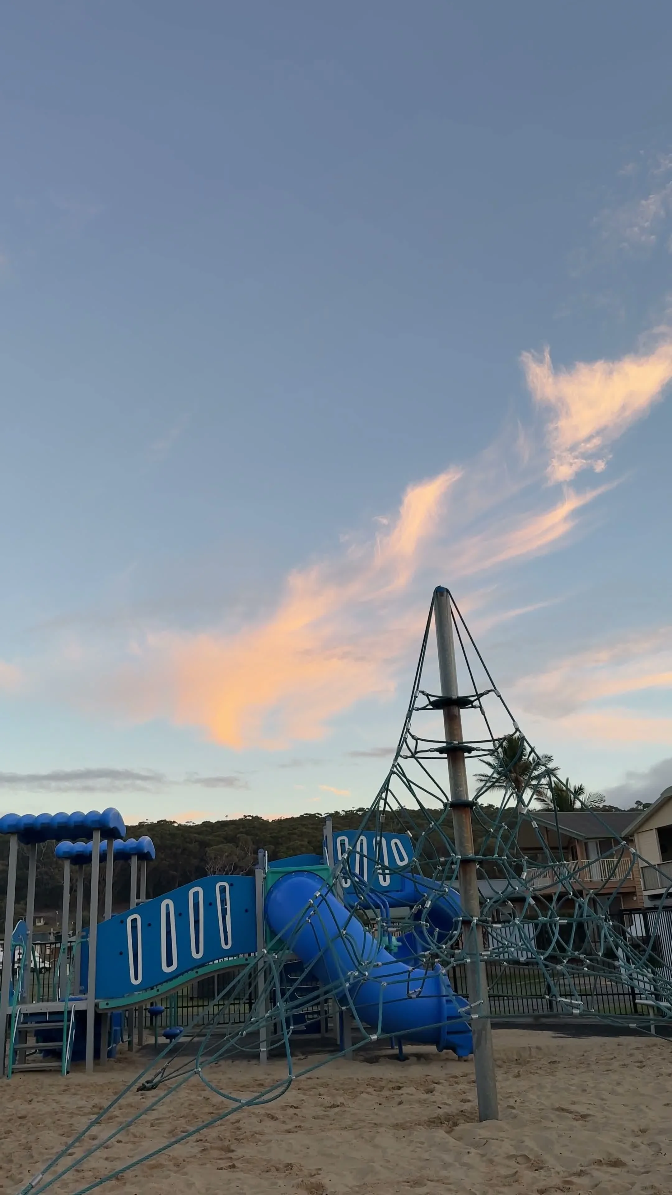 An outdoor playground with blue slides and climbing structures, set on sandy ground, with a sky showing clouds during sunset in the background.