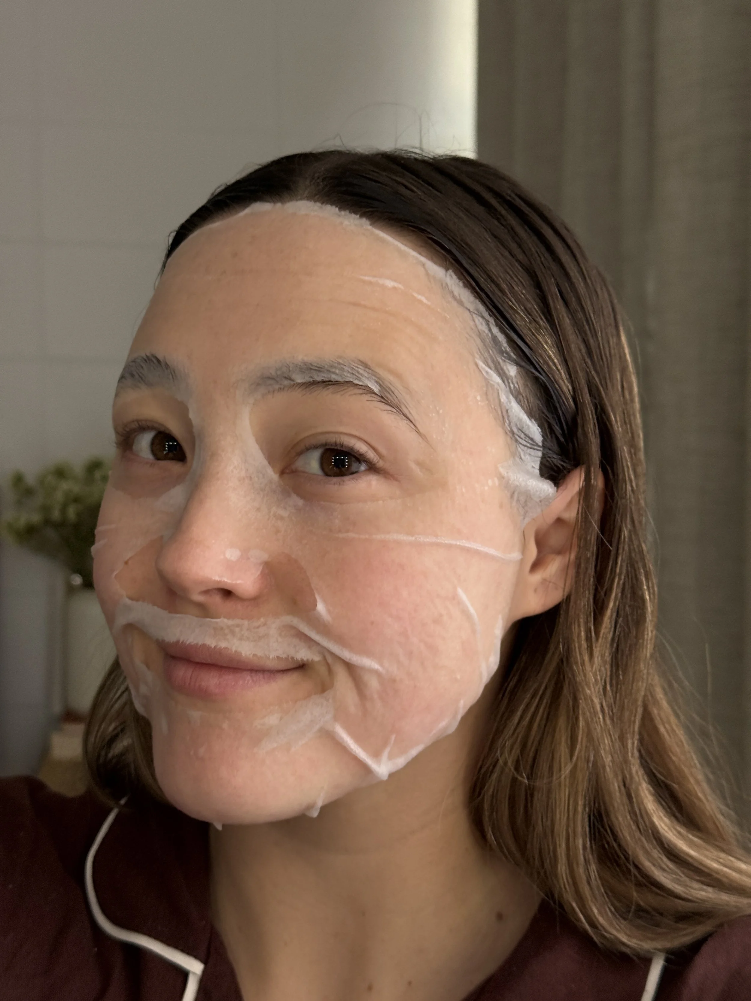 Close-up of a woman with long brown hair, smiling slightly, with a facial sheet mask applied to her face, partially visible at the chin and cheeks.