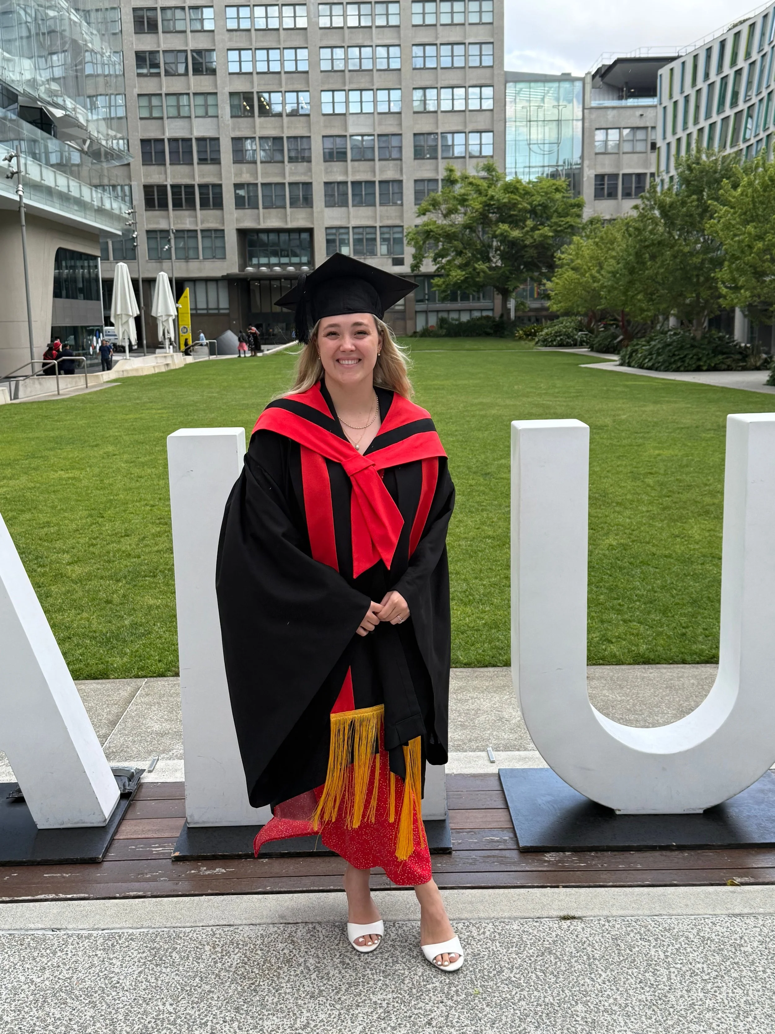 A woman in a graduation gown and cap stands outdoors in front of large white letters spelling 'U' and part of 'N'. She is smiling, dressed in a red dress with black floral patterns and white open-toe shoes. The background shows a green lawn, modern office buildings, and a few people walking.