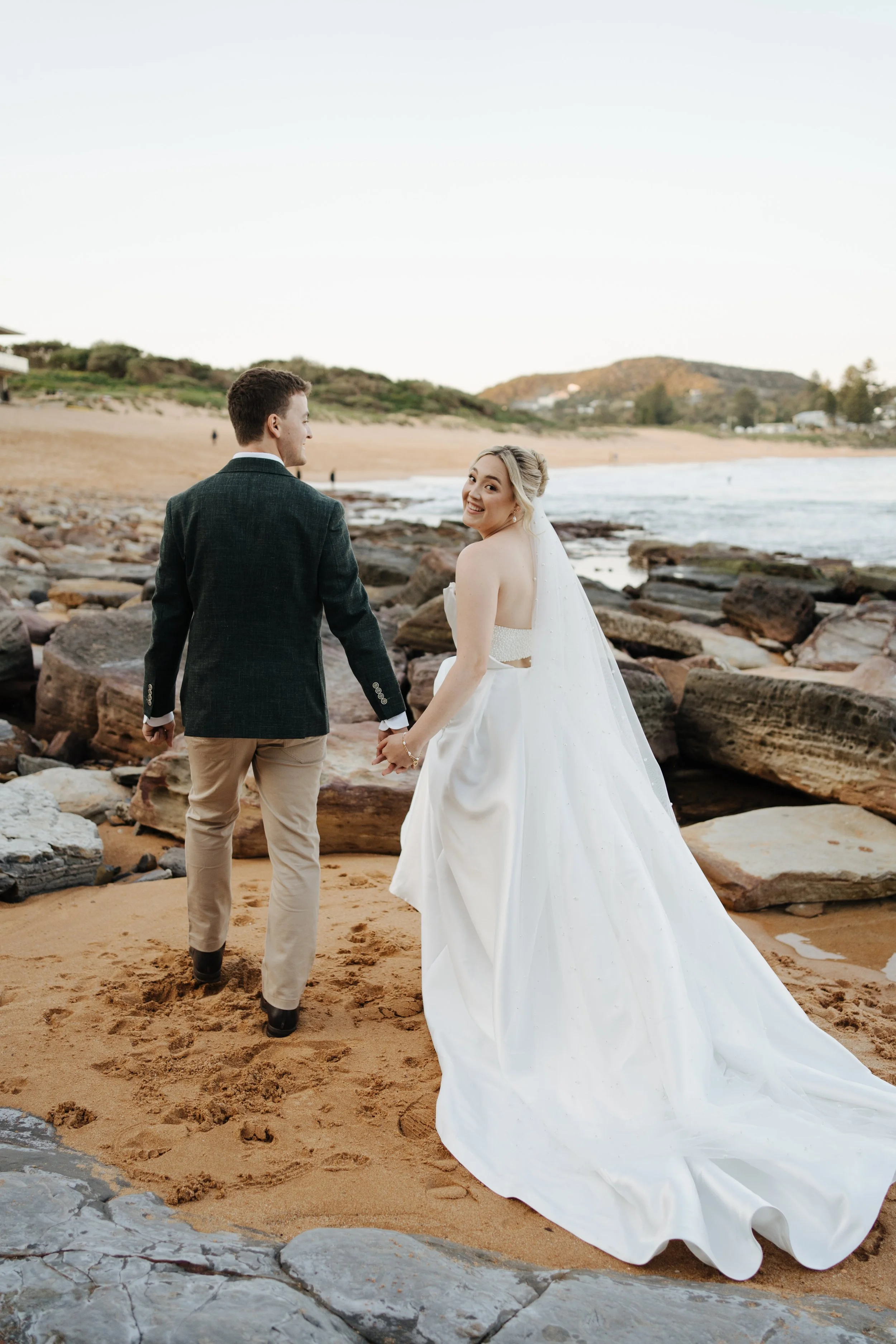 A bride and groom holding hands on a beach, with the bride smiling back at the camera, wearing a white wedding gown and veil, and the groom in a dark blazer and khaki pants, with rocks and sand in the foreground and hills in the background under a clear sky.