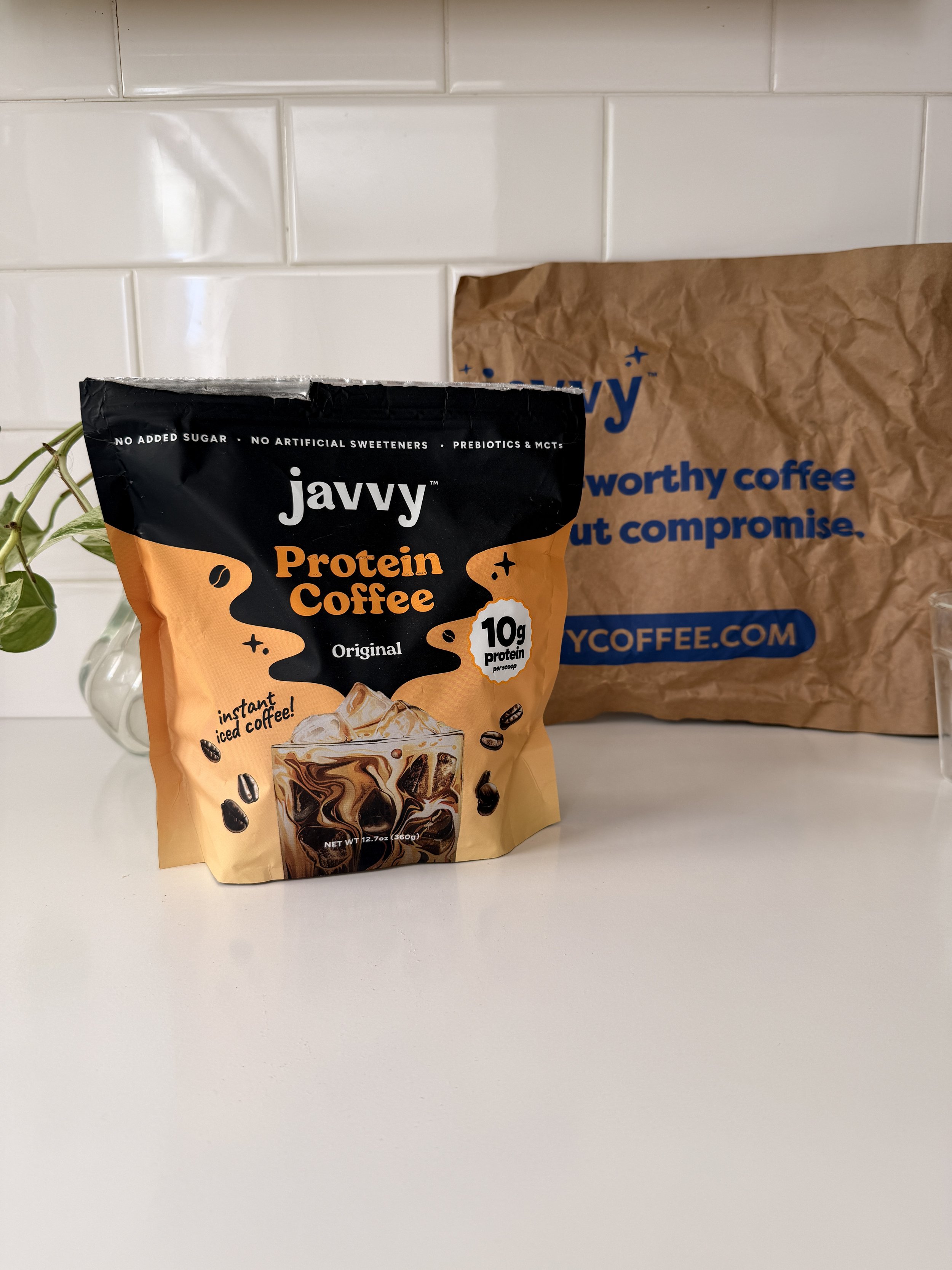 A bag of Javvy Protein Coffee on a white surface with a paper bag behind it and a small plant to the left.