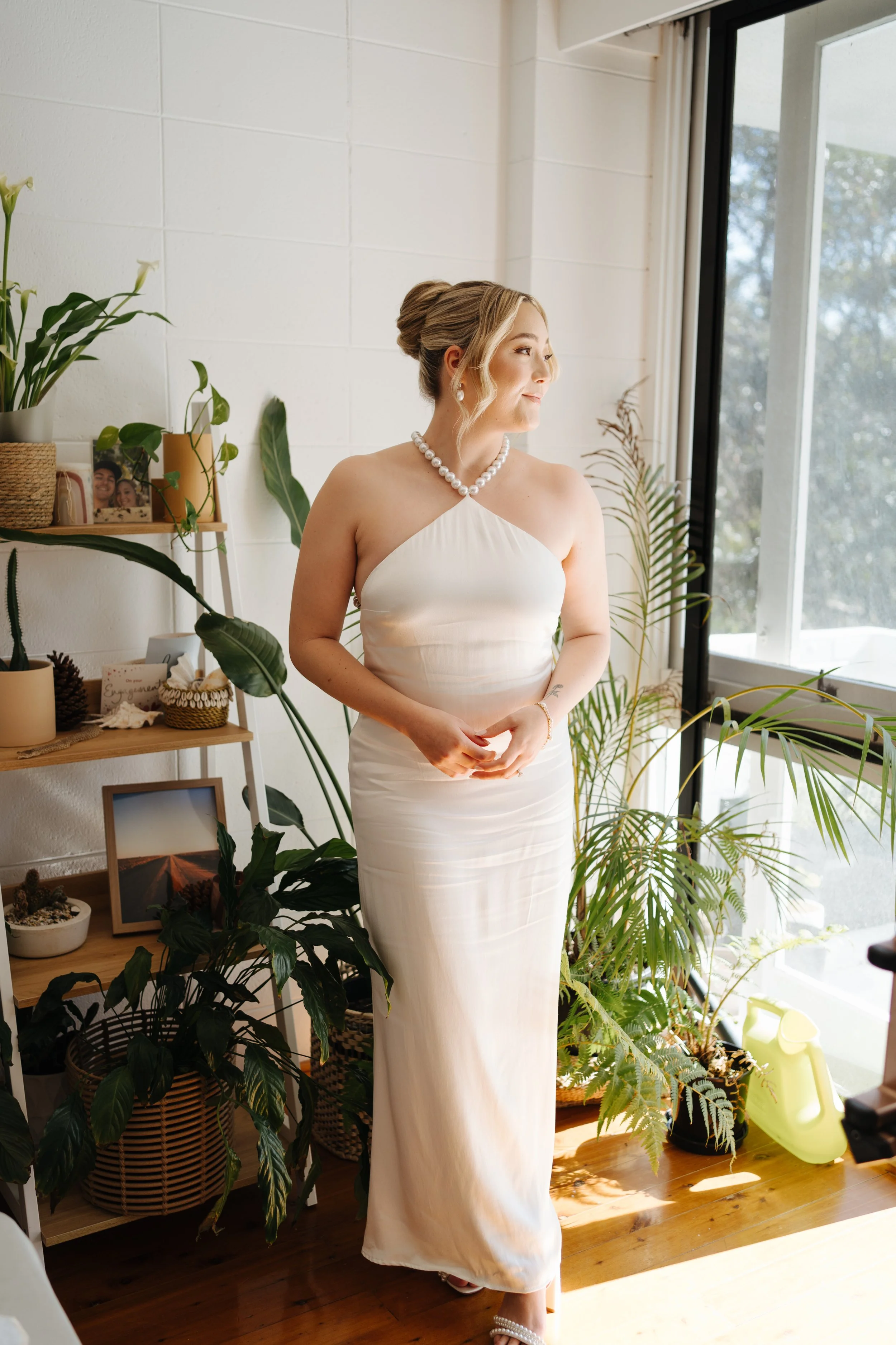 A woman in a white strapless dress standing near large windows with sunlight and surrounded by potted plants, smiling and looking to the side.