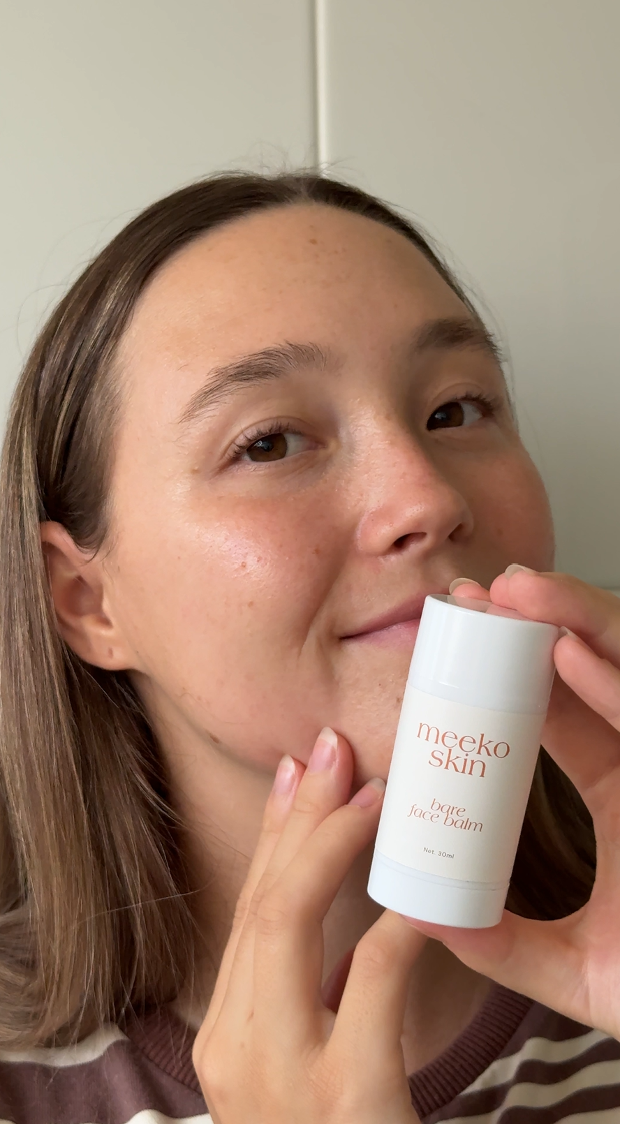 A young woman with fair skin and brown hair holding a white bottle of Meeko Skin Bare Face Balm close to her face, smiling softly, in front of a plain white wall.