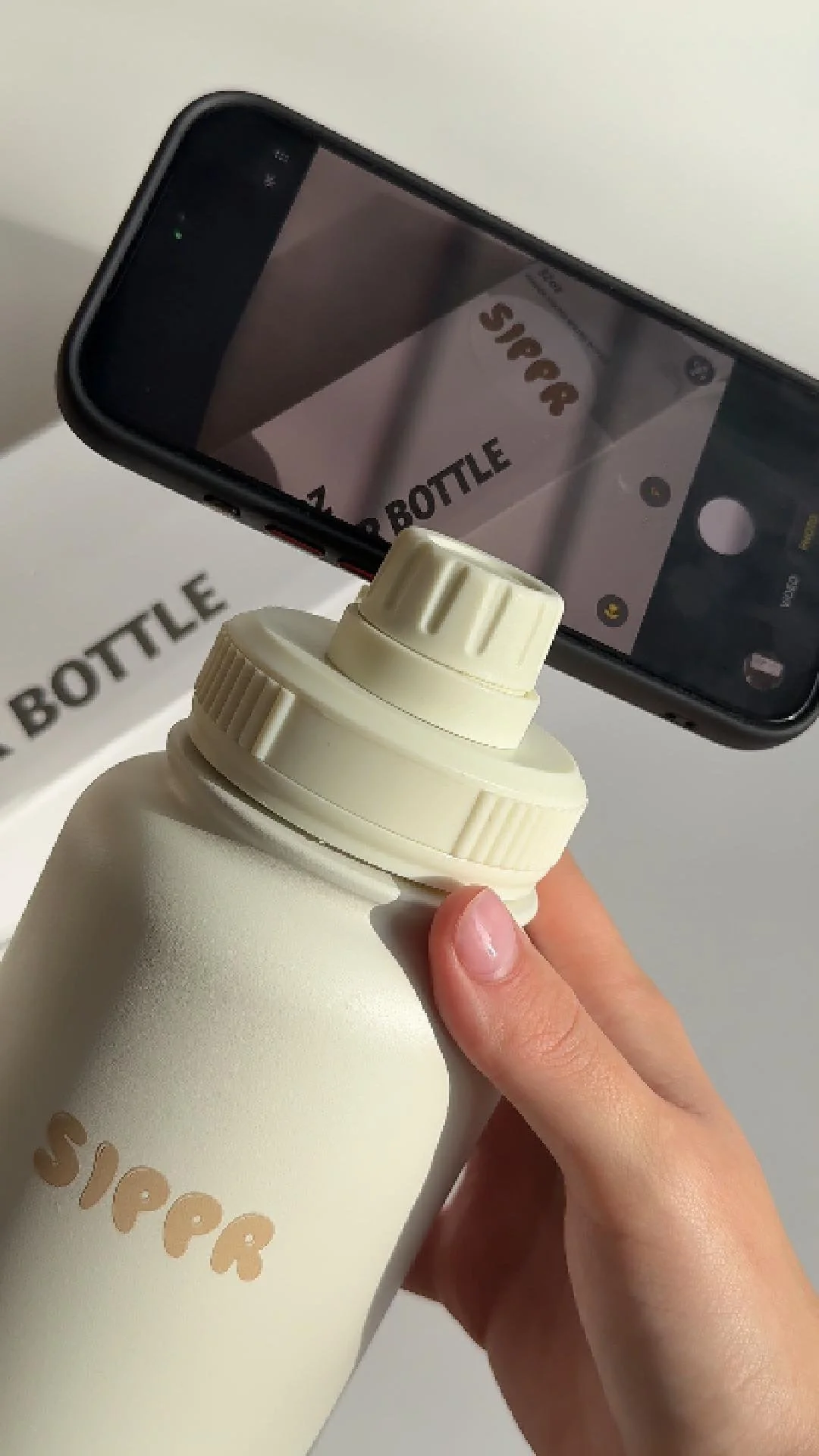 Person holding a white water bottle with a screw-on lid, camera capturing the water bottle and a phone taking a picture of the bottle, background shows a box with the word 'Super' on it.