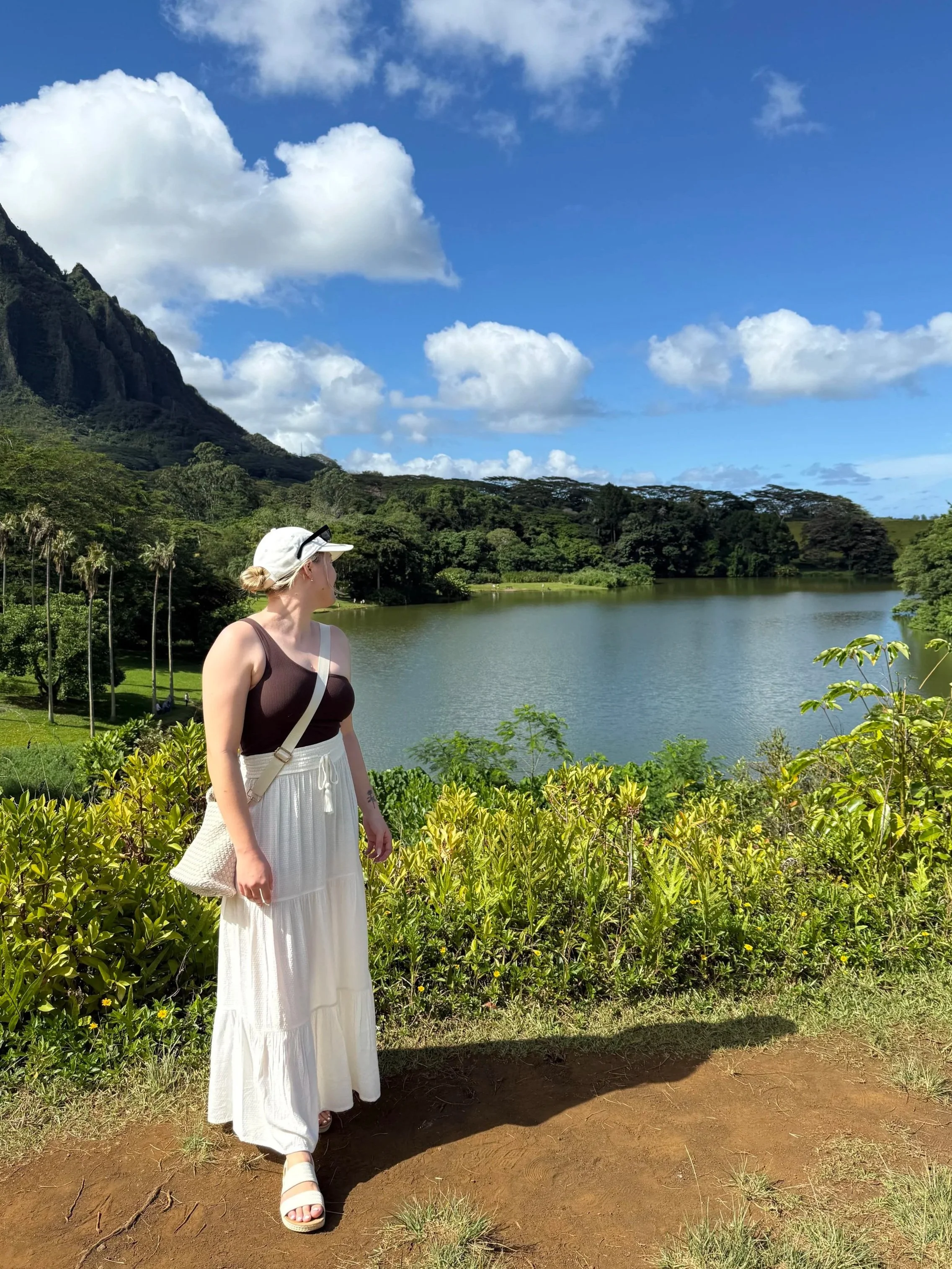 A woman with blonde hair in a bun, wearing a white hat, sunglasses, a dark tank top, and a long white skirt, standing outdoors near a body of water with a mountainous landscape, lush greenery, blue sky, and scattered clouds in the background.